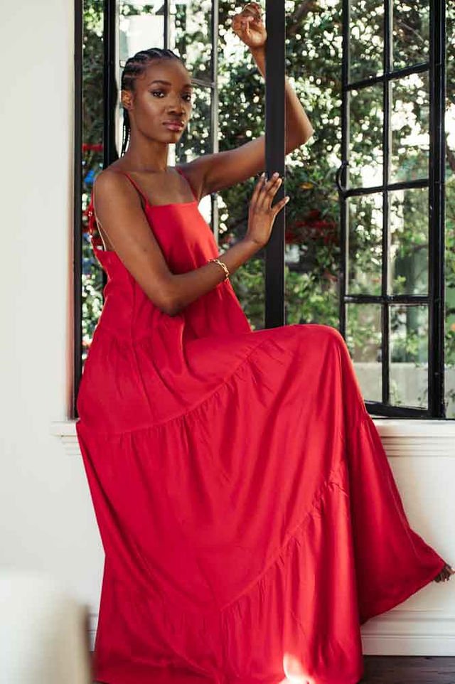 In a luxurious brand photoshoot, a woman wearing a red dress gracefully poses on a window sill.