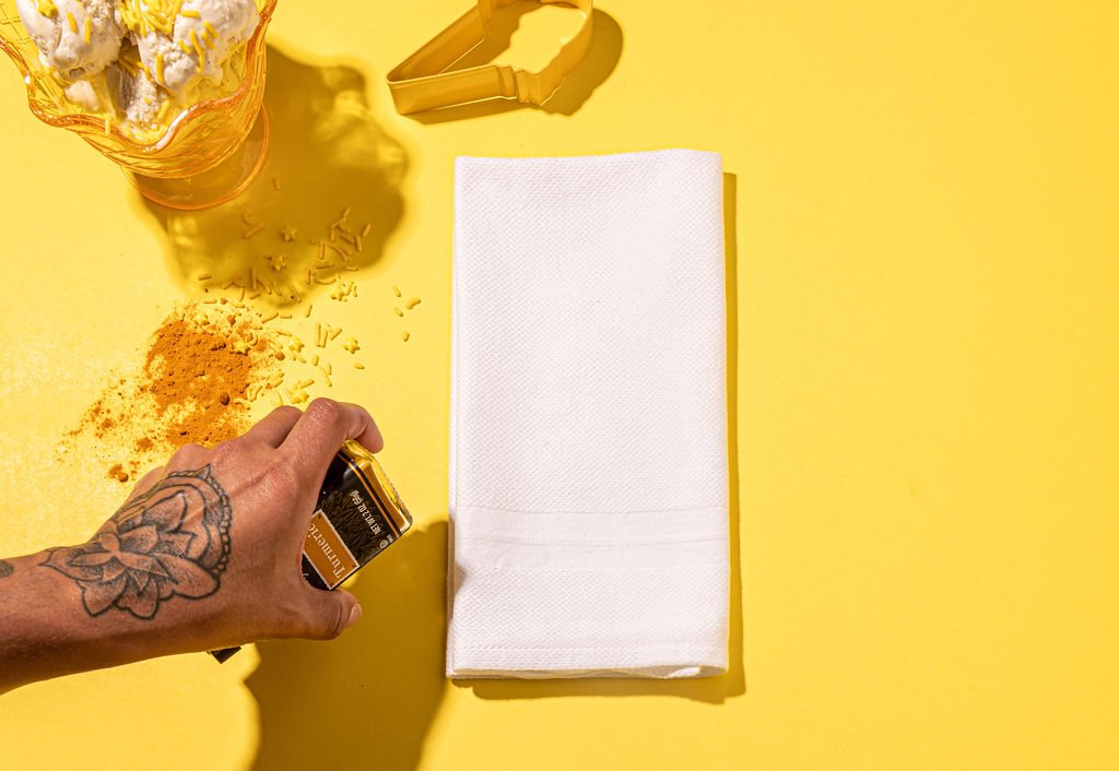 A person with tattoos holding a white napkin made of Cotton Linen over a vibrant yellow surface.