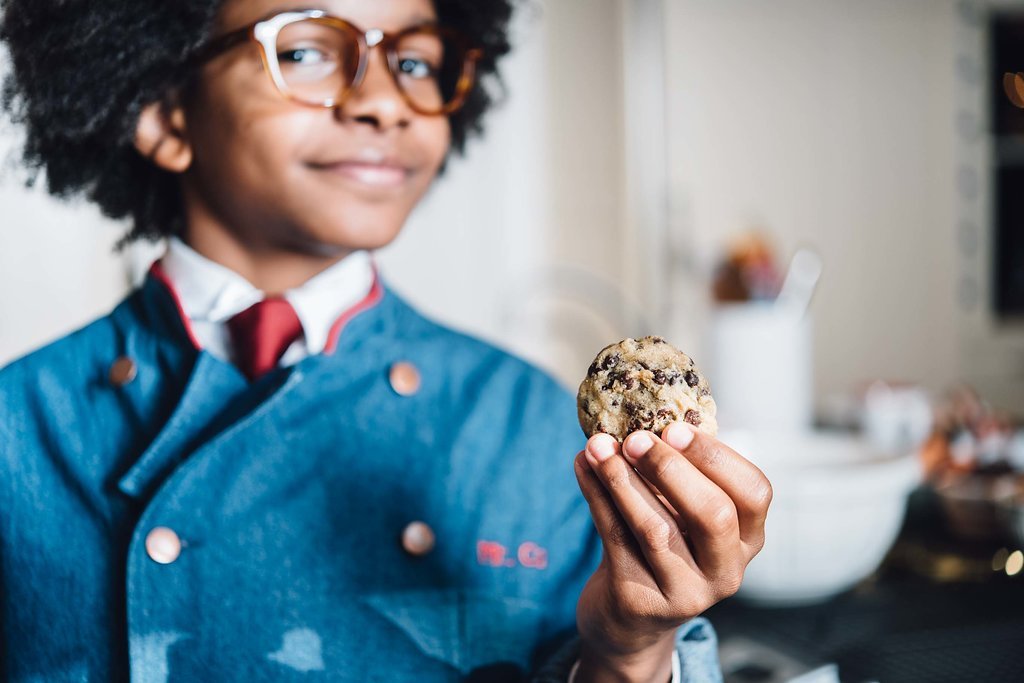 MR CORY'S COOKIES, in personal branding photography, presents a young boy in a blue jacket holding a delicious cookie.