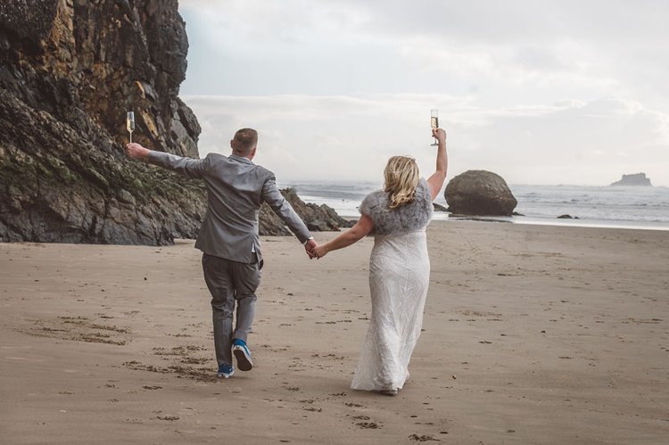 Newlyweds holding hands, walking on the beach with champagne glass in their hands.jpg
