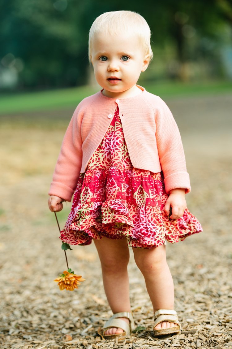 A family photographer captures a baby girl in a pink dress and jacket standing on a dirt road.jpg