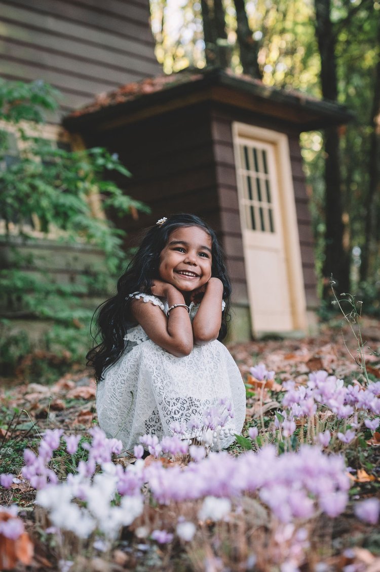 A little girl in a white dress sits amidst flowers in front of a cabin, creating a quaint scene.jpg