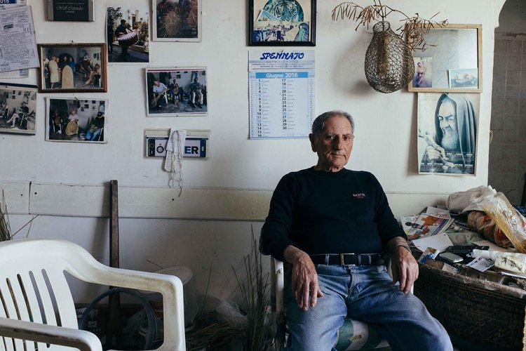 An elderly man sits in a room with a wall of pictures. A travel photographer captured this image during his explorations.jpg