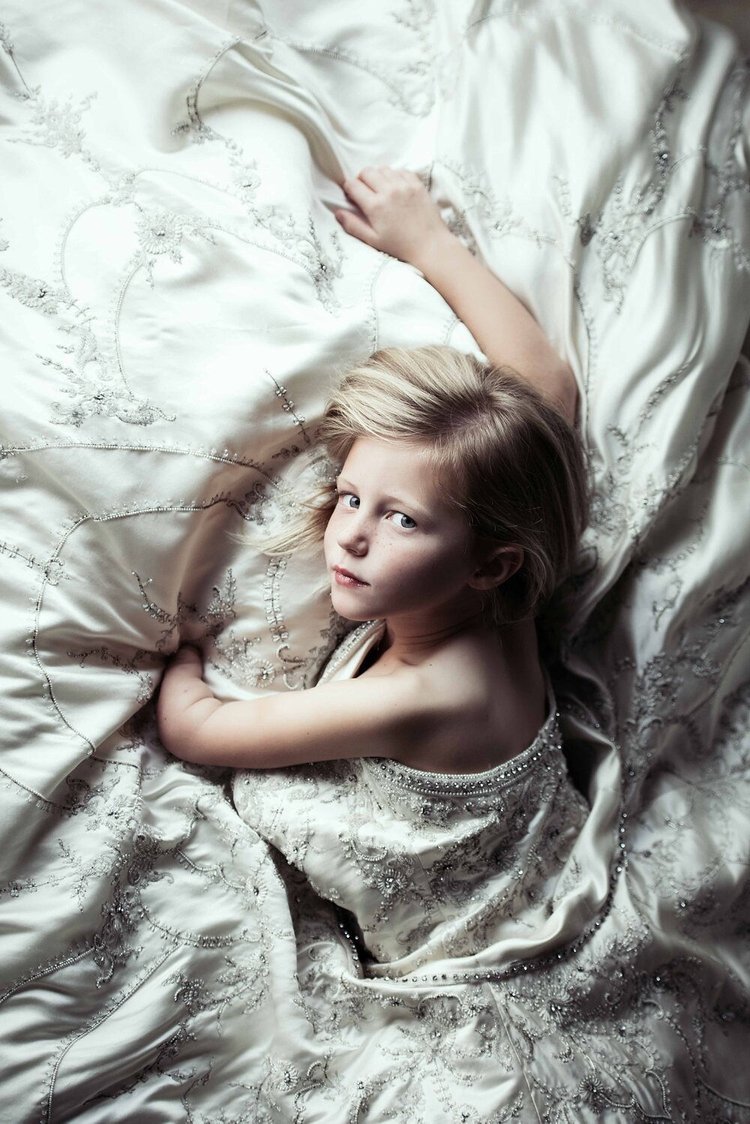 A Portland children's  photograph captures a young girl in an adorned white dress, peacefully lying on a bed.jpg