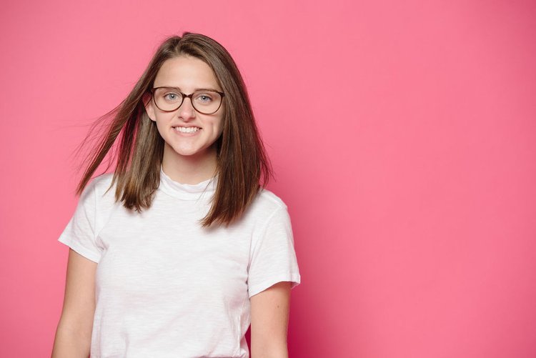 A young girl with glasses and a white t-shirt, posing against a pink background for Children Photography.jpg