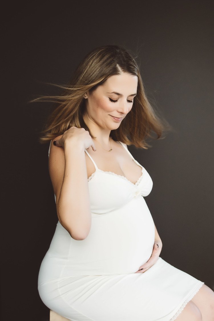 An elegant pregnant woman in a white dress, posing on a stool, photographed by a lifestyle family photographer.jpg