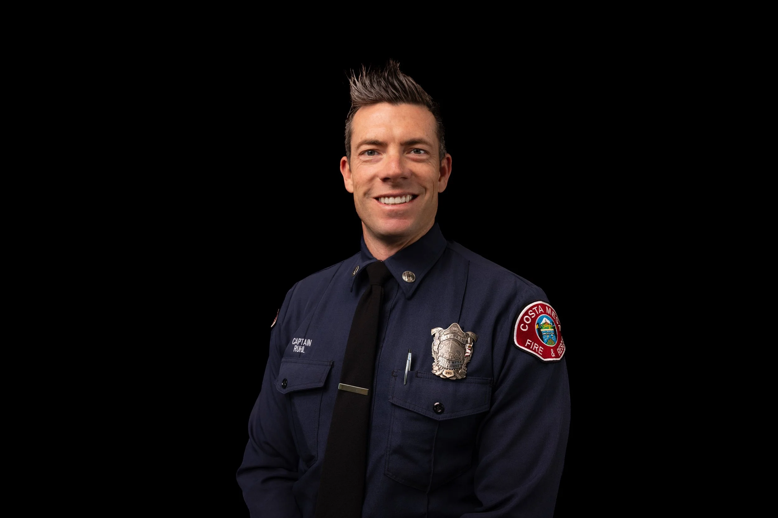 A portrait of a smiling male police officer with short dark hair and blue eyes, wearing a dark police uniform with badges, against a plain dark background.