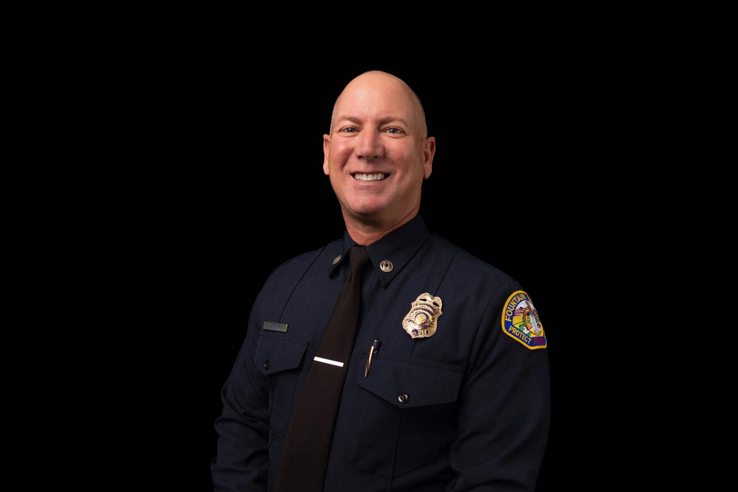 A male firefighter in uniform, smiling, with a badge and an embroidered patch on his sleeve, against a black background.