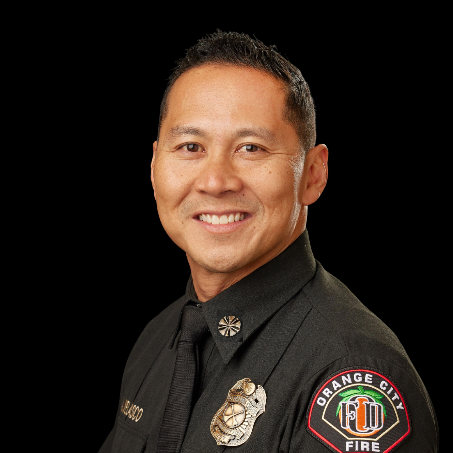 Portrait of a smiling man in a black uniform with a cross pin on the collar and a badge on the sleeve, against a plain gray background.