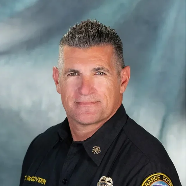 A middle-aged man with short, dark hair and light skin worn in a professional uniform, featuring a badge and a patch that reads 'Orange County.' He is smiling slightly against a blurred, cloudy background.