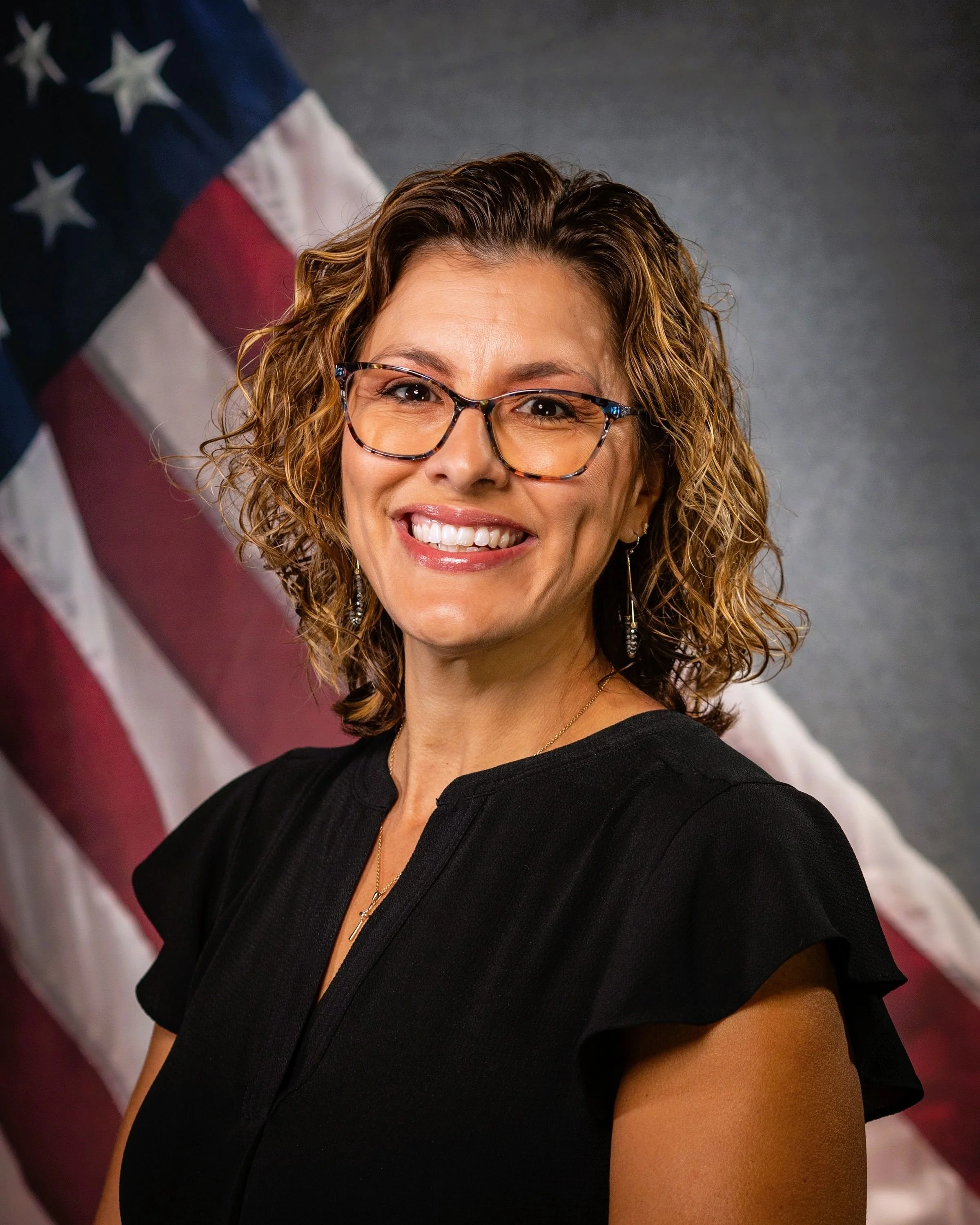 A woman with curly brown hair, wearing glasses and a black dress, smiling in front of a partially visible American flag.