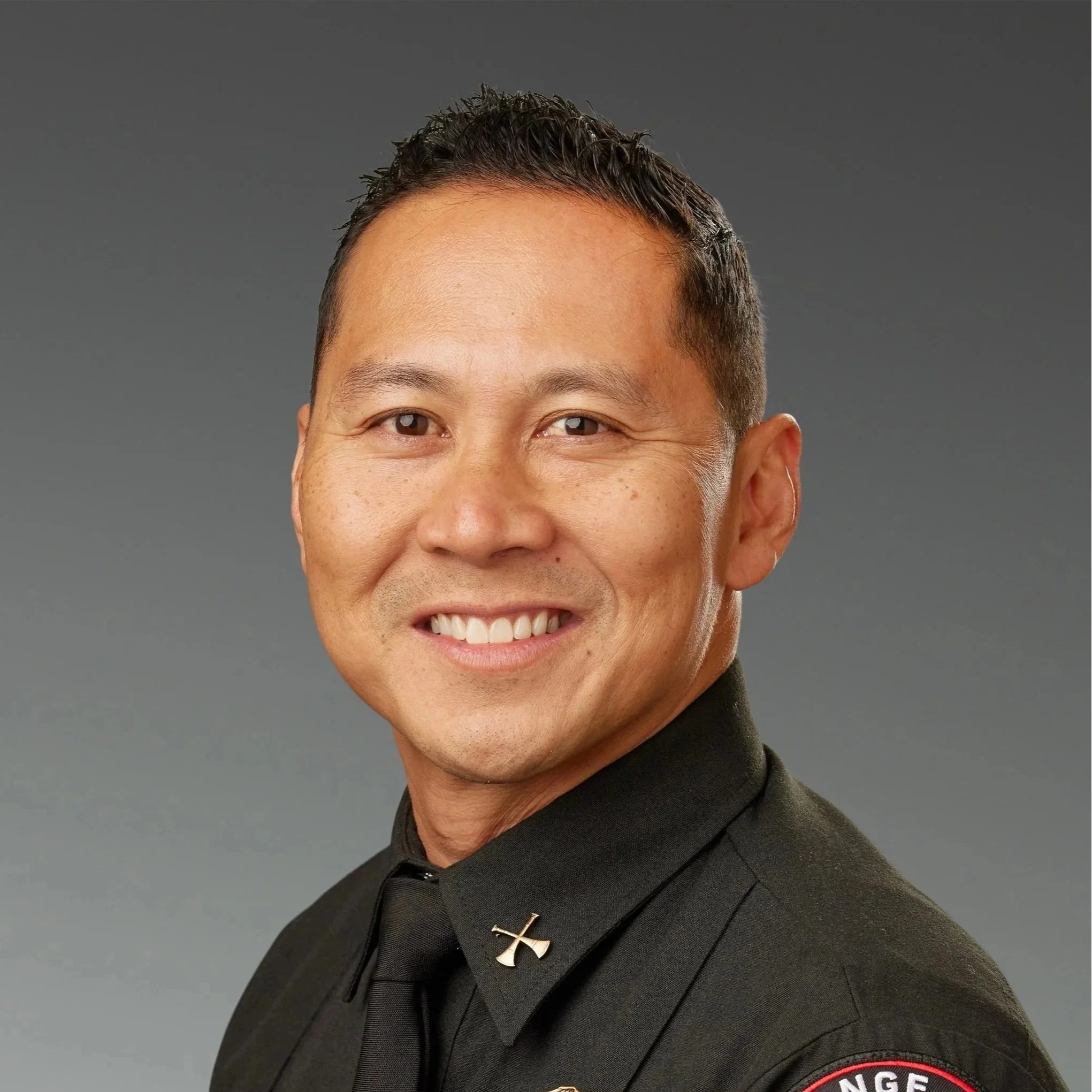 Portrait of a smiling man in a black uniform with a cross pin on the collar and a badge on the sleeve, against a plain gray background.