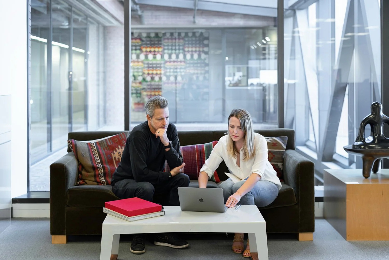 Two people sitting on a black sofa looking at a laptop in an office lounge with large windows and modern decor.