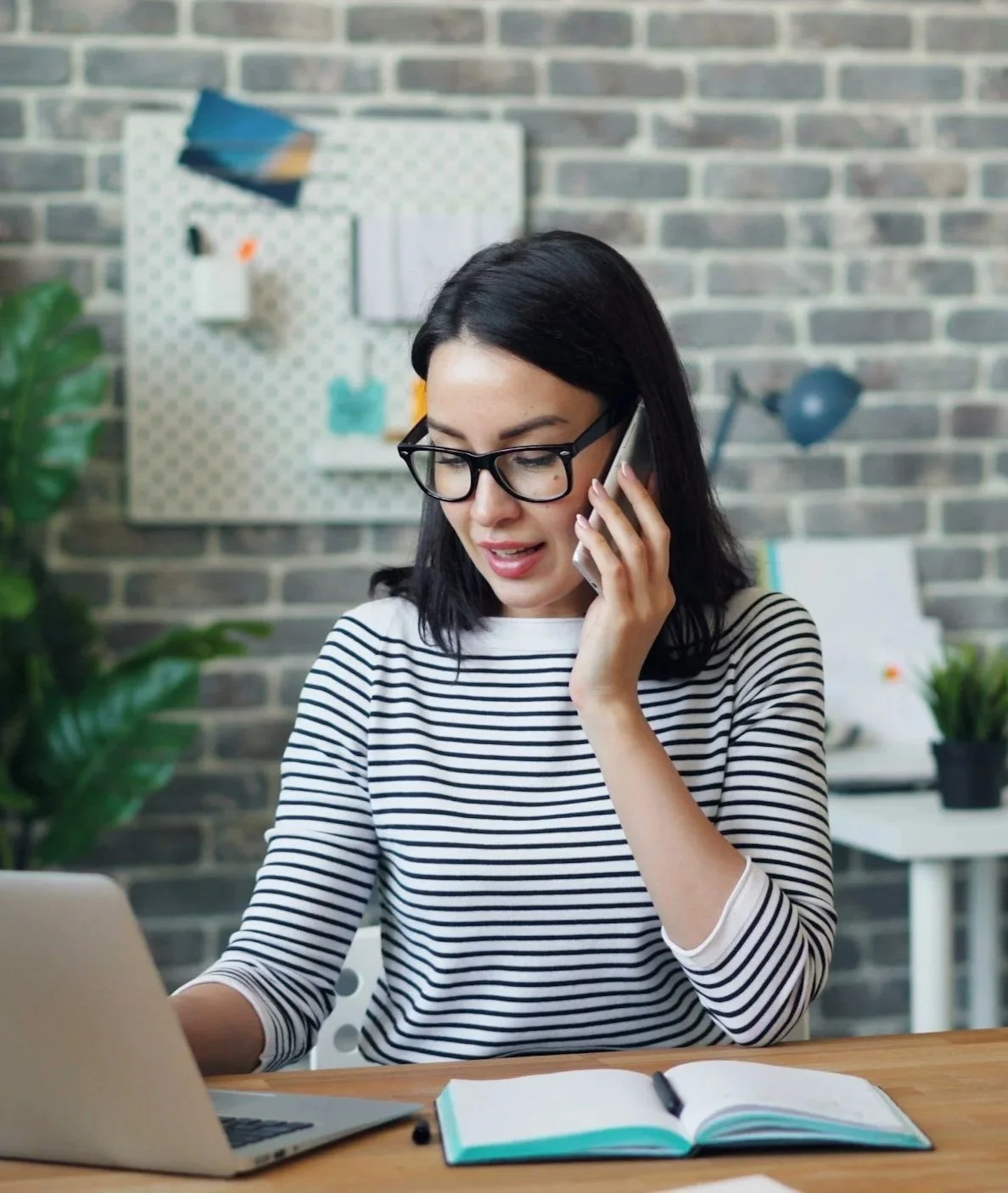 A woman with black hair and glasses, wearing a striped shirt, talking on the phone while working at a desk with a laptop and an open notebook in a modern office setting.