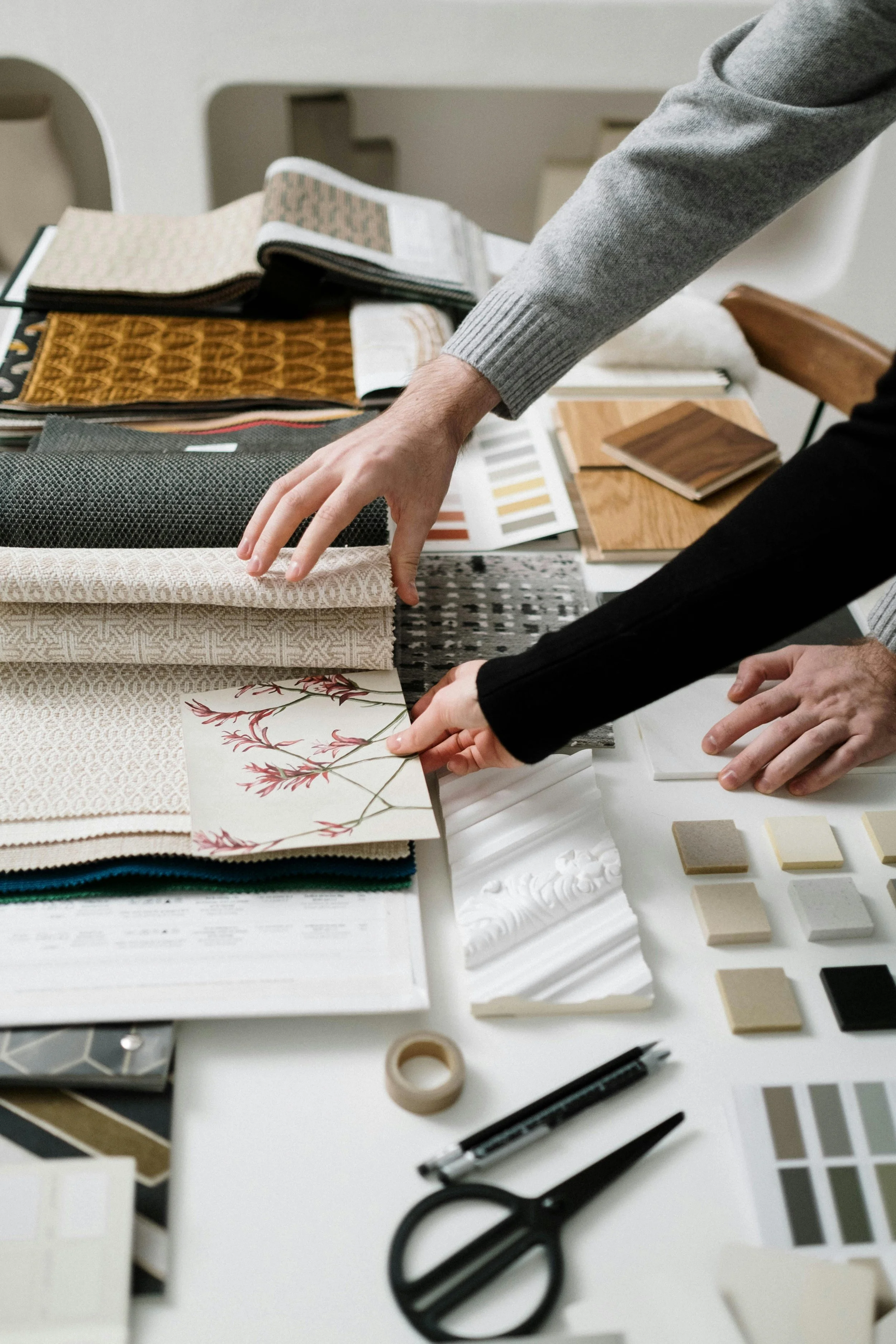 Two people are selecting and examining fabric and material swatches on a table, which also contains color samples, paint chips, scissors, a pen, and decorative molding samples, likely for interior design or renovation planning.