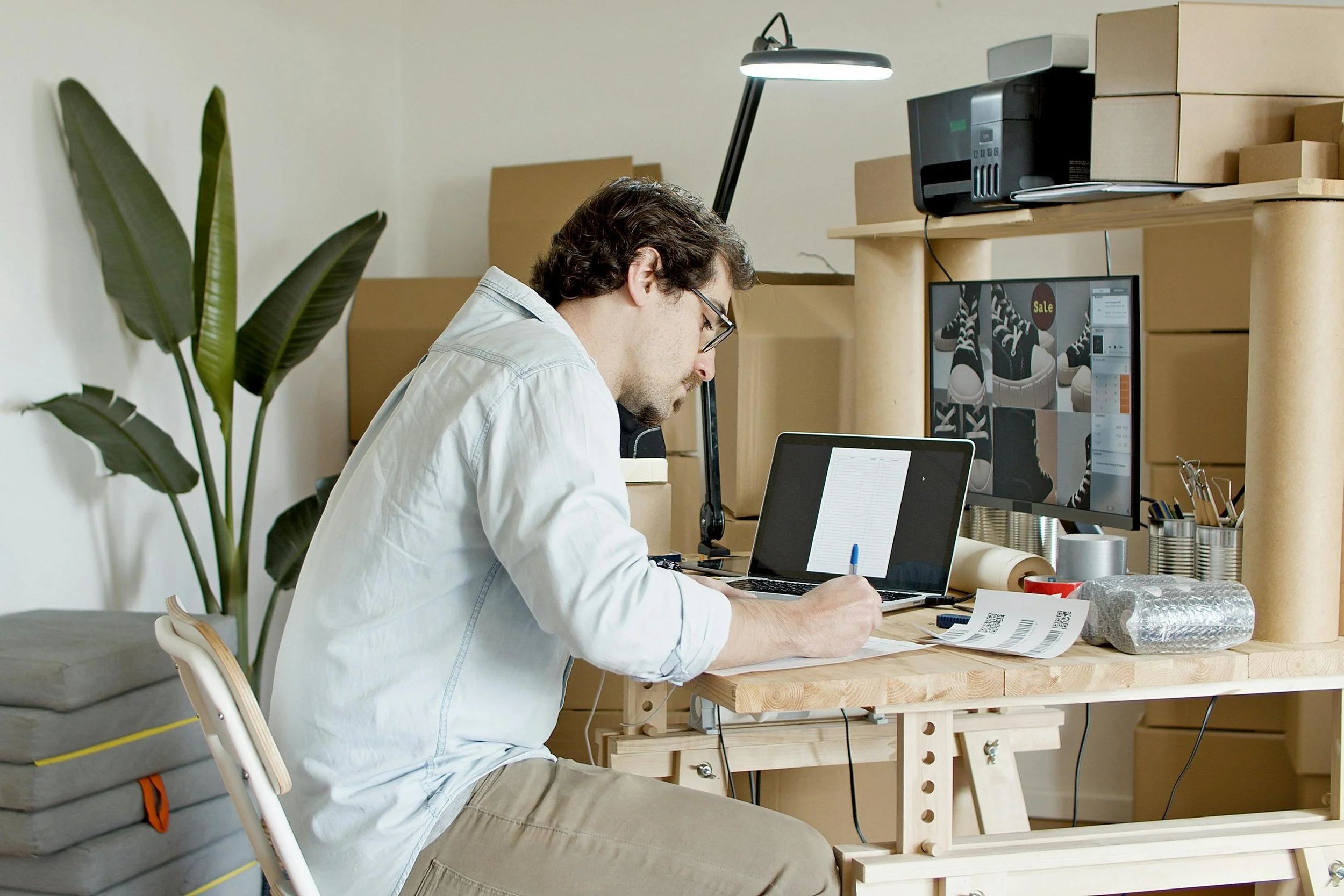 Man working at a wooden desk, writing on paper with a pen, surrounded by packing materials, a laptop, and a computer monitor showing shoe images.