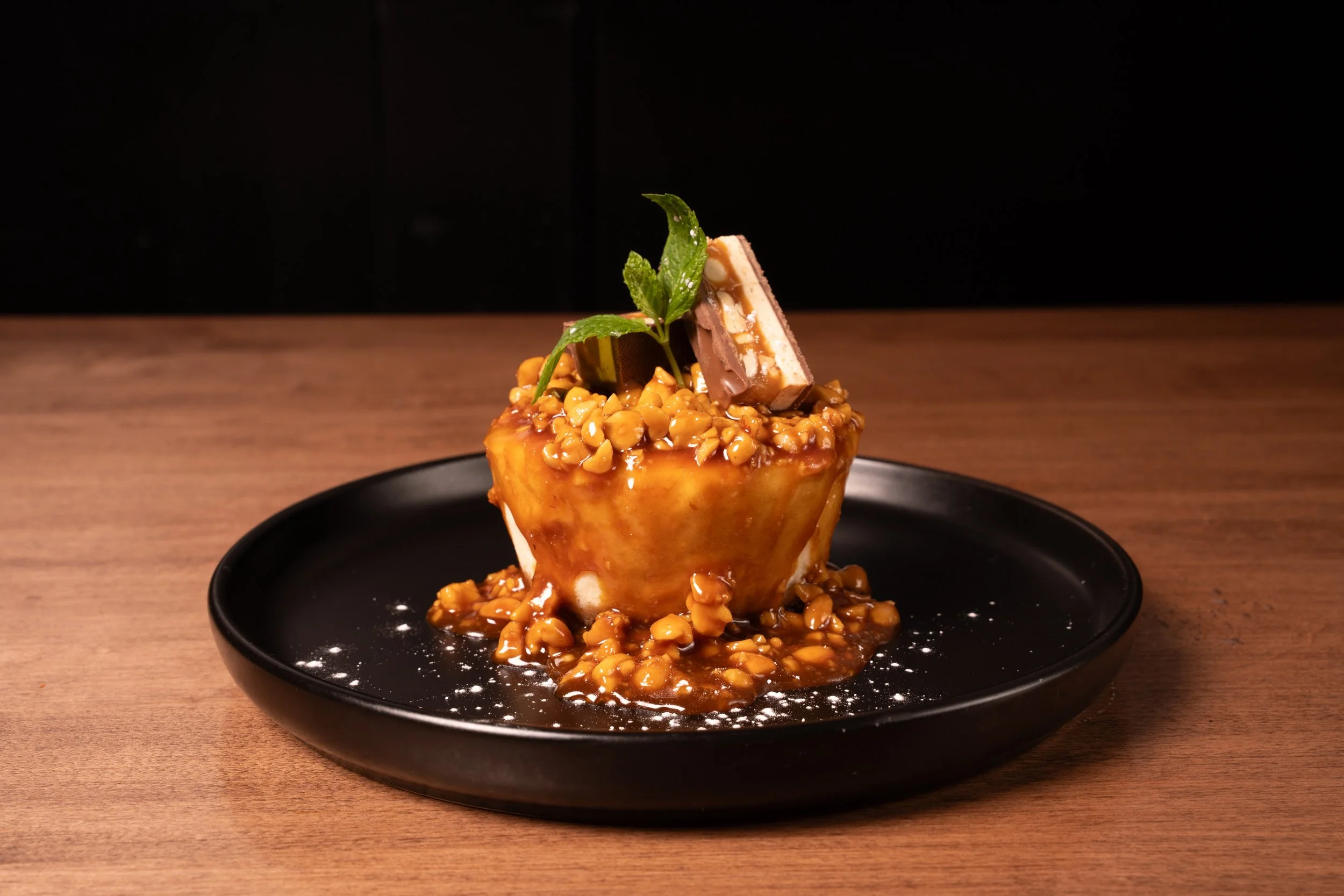Salted Caramel Snickers Cheesecake: house-made cheesecake topped with salted caramel, snickers, roasted peanuts, chocolate and powdered suger.  Dessert Brix + Barrel.  