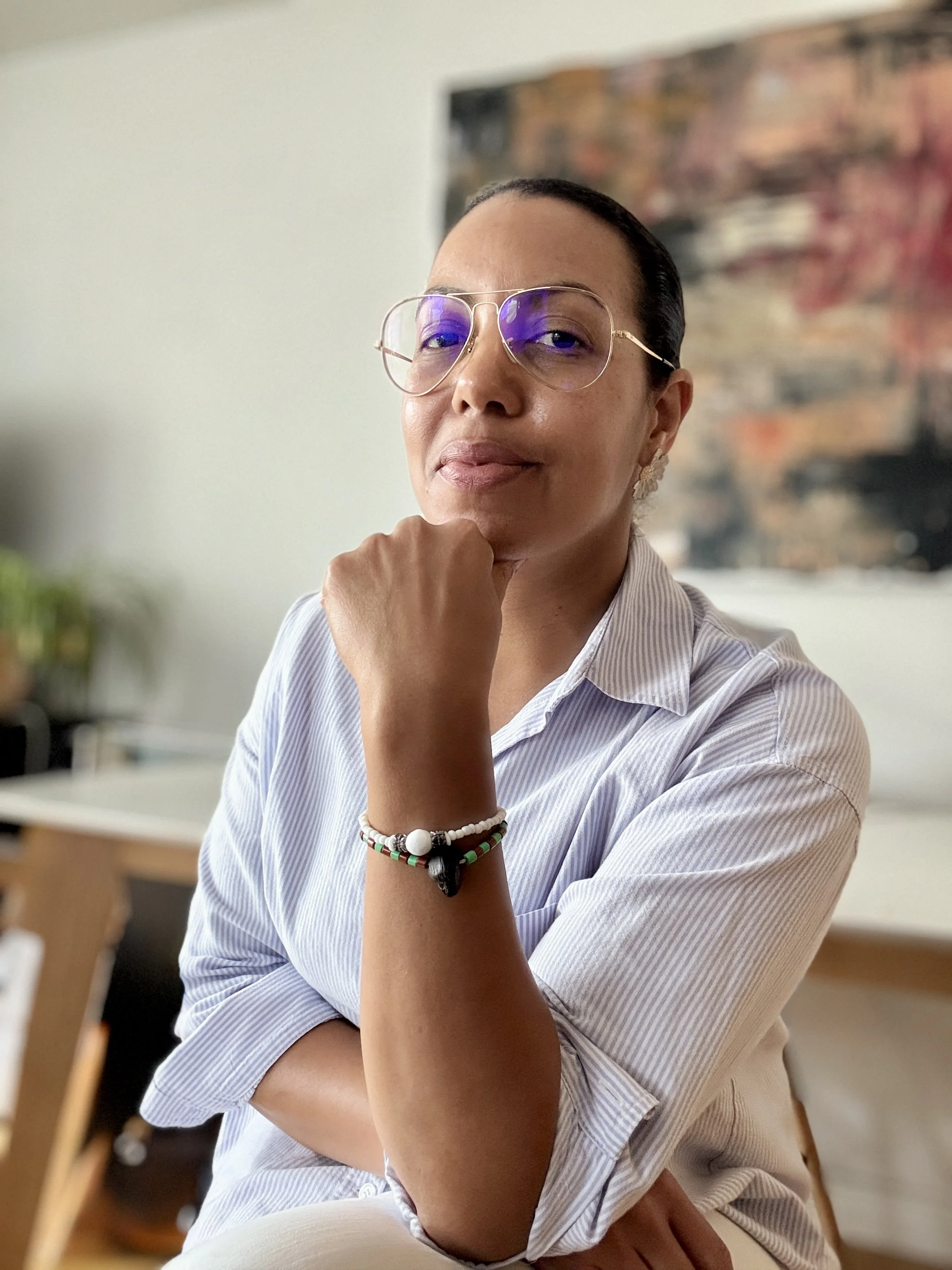 Person wearing blue-tinted glasses and a striped shirt, sitting at a desk with natural light, looking directly at camera with a thoughtful expression