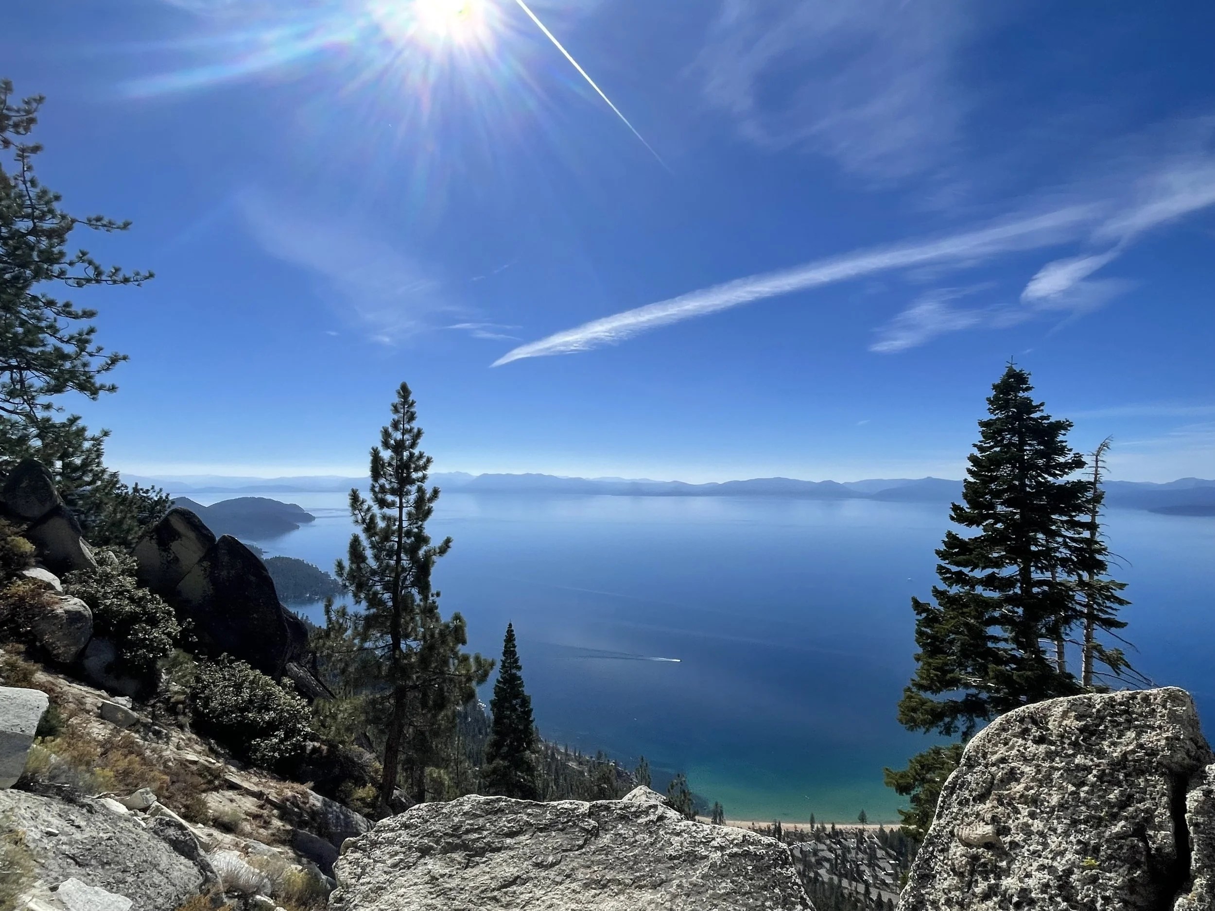 A scenic view of a large blue lake surrounded by forested mountains, with tall pine trees in the foreground, large rocks, and the sun shining brightly in a clear blue sky.