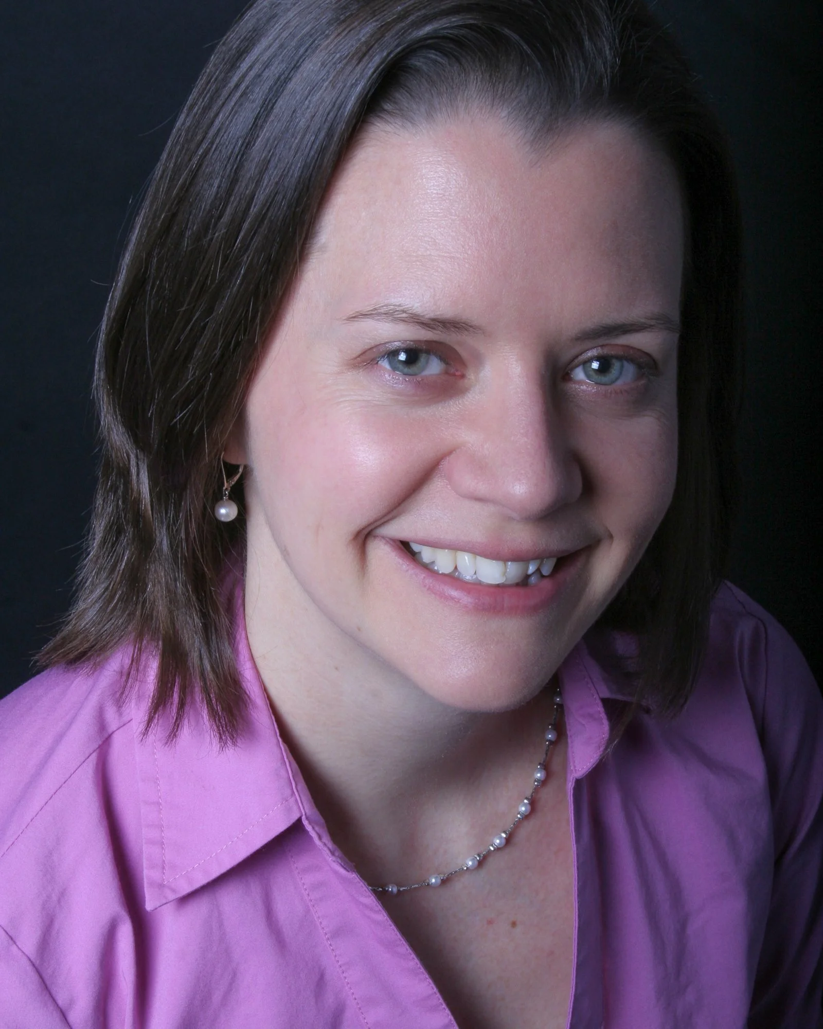 Close-up of a woman smiling, with blue eyes and brown hair wearing a purple shirt, pearl earrings, and a pearl necklace against a black background.