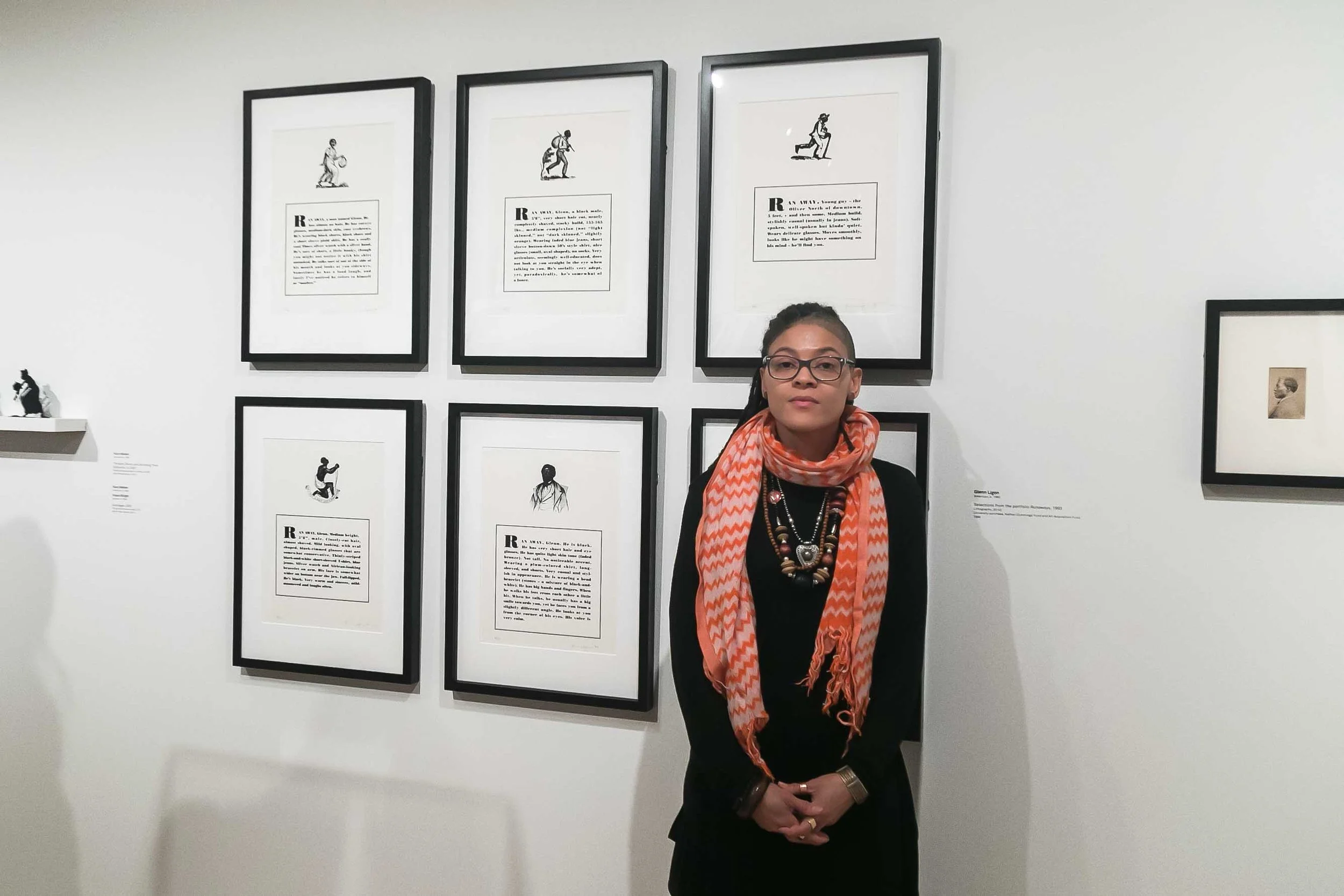 A woman with glasses and a patterned orange scarf stands in front of framed artwork and read text, likely in a gallery or museum.