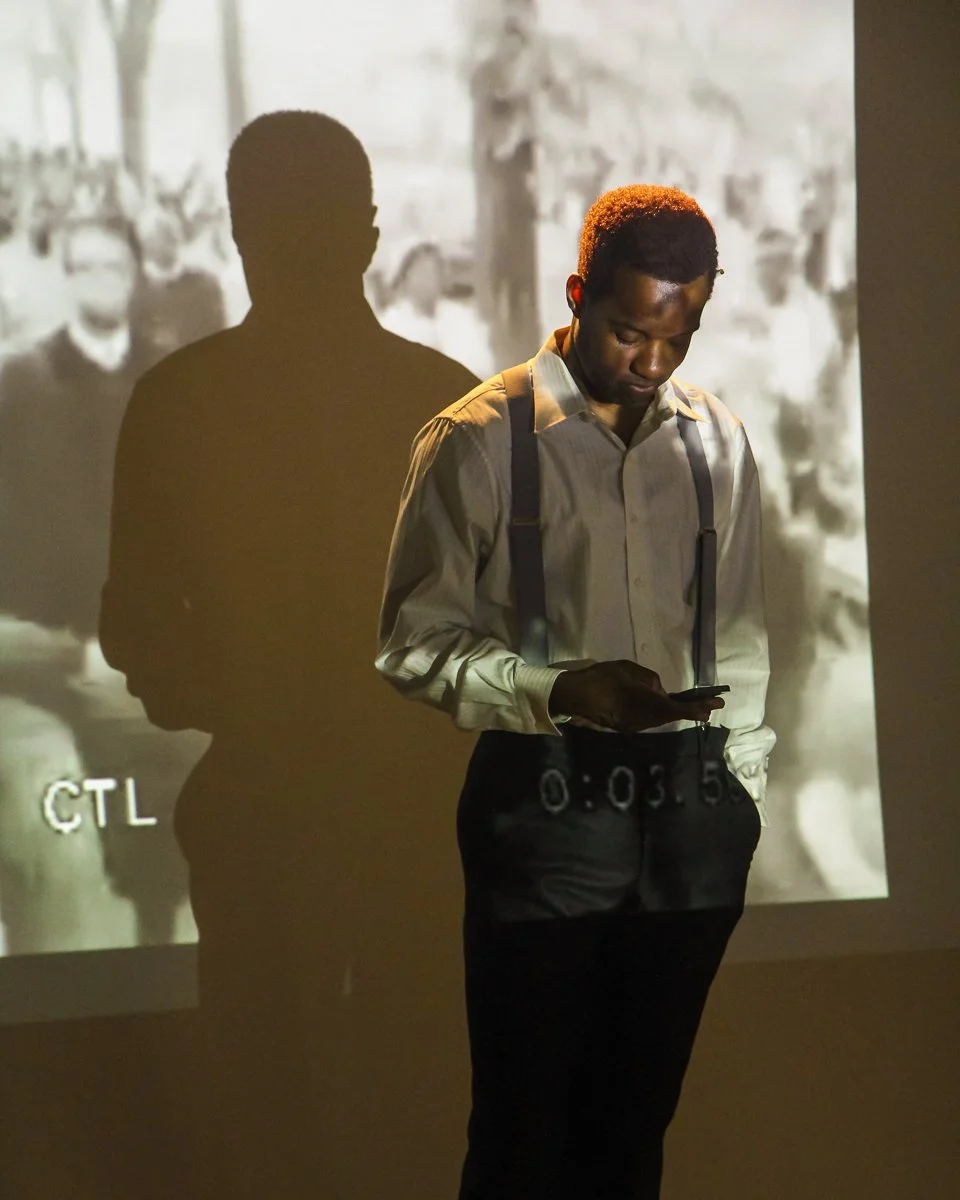 A man in a light-colored shirt with suspenders stands indoors, looking at his phone. His shadow is cast on a wall behind him, where a black-and-white projection shows a group of people, possibly from a historical photo.