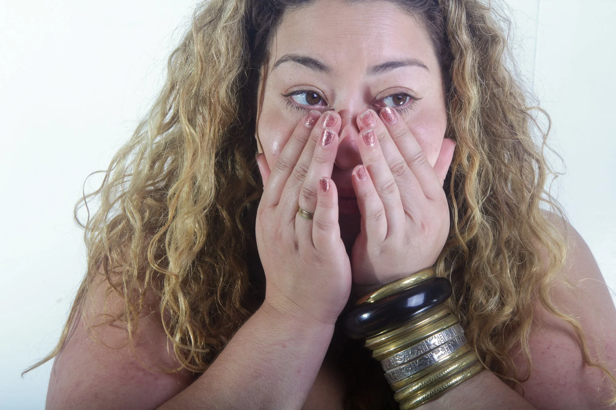 A woman with curly blonde hair covering her nose with both hands. She is wearing multiple gold and silver bangles on her right arm and has glittery pink nail polish.