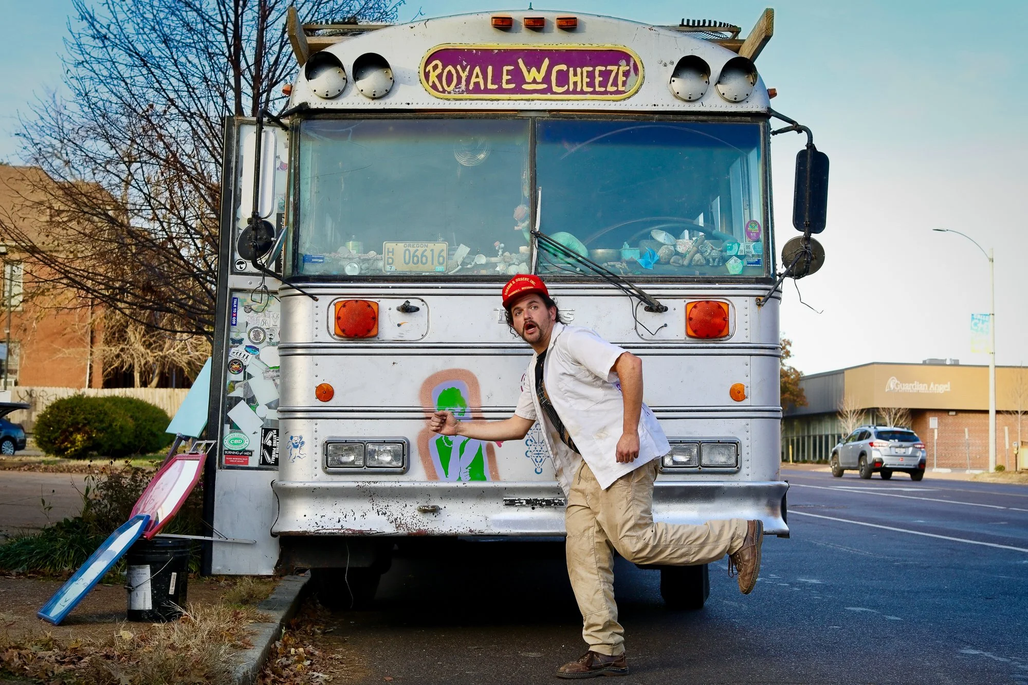 A man with a mustache, wearing a red cap, white shirt, and tan pants, is leaning on an old food truck labeled 'Royal Cheeze,' with the man holding something in his hand. The truck's windshield is cluttered with items, and the truck is parked near a s