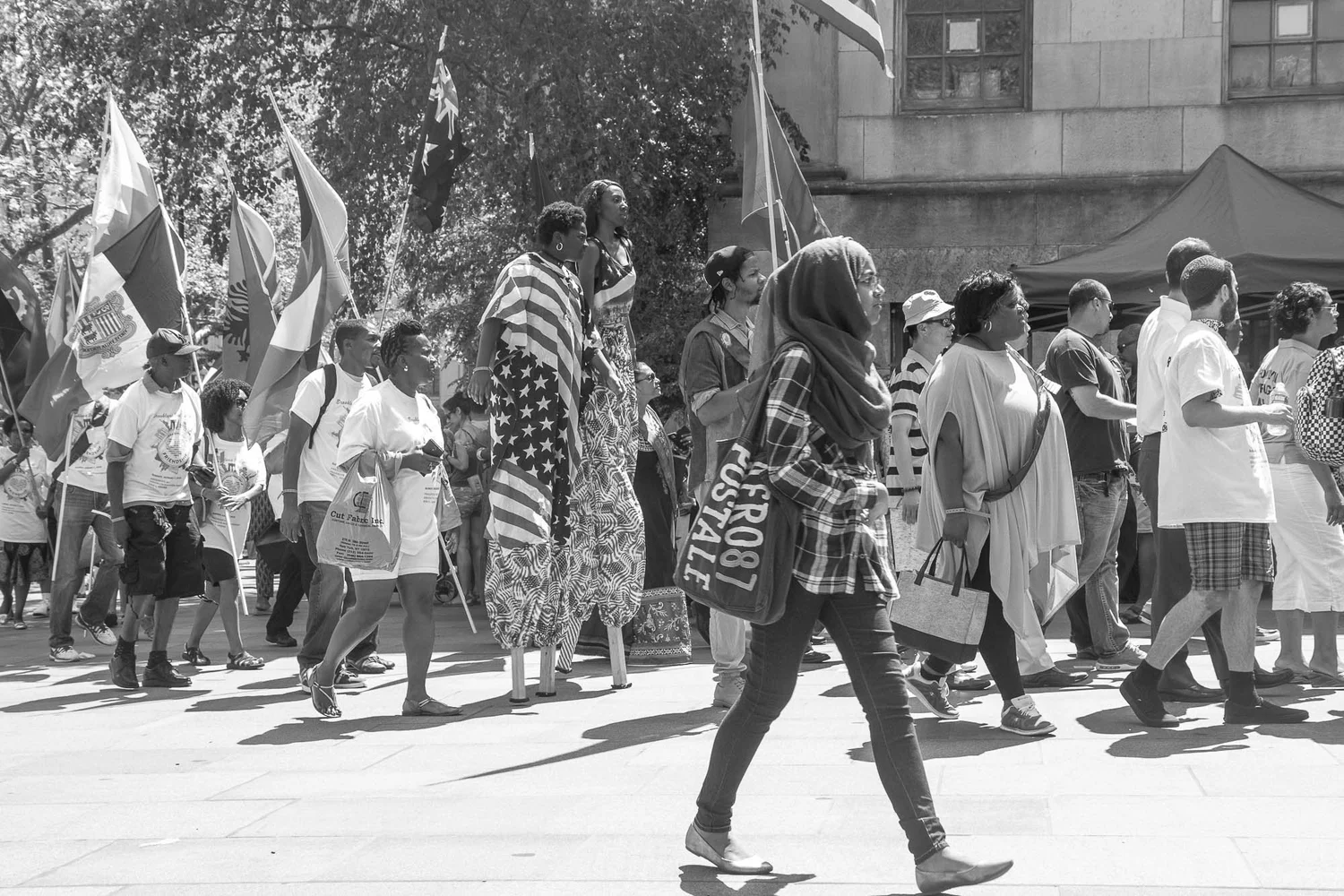 Black-and-white street scene with a passing crowd, flags, and two people on stilts.