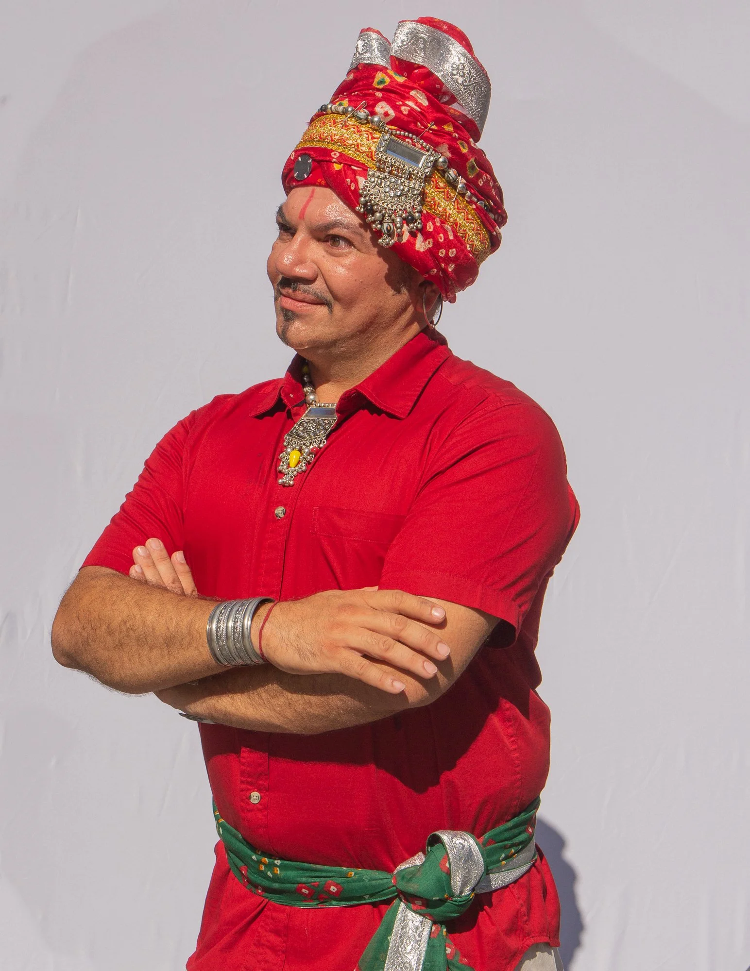 A man wearing a colorful traditional Indian outfit, including a red shirt, a green and red sash with silver accents, and a large intricately decorated headwrap. He has his arms crossed and is standing against a plain light-colored background.