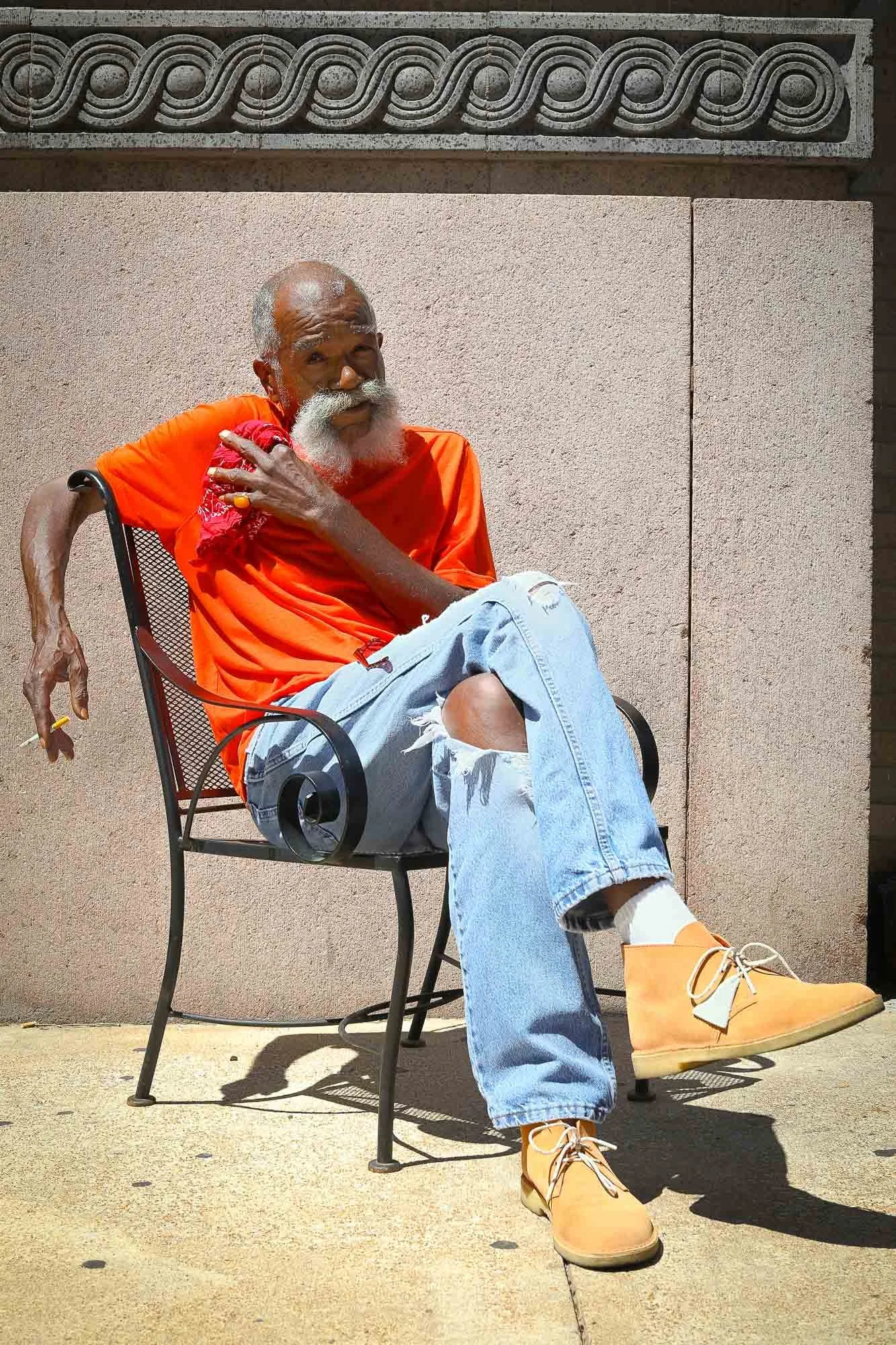 An elderly man with a white beard and bald head sitting outdoors on a metal chair, wearing an orange shirt, ripped jeans, and tan boots. He has one hand on his shoulder and is looking upward. The background depicts a beige wall with a decorative conc