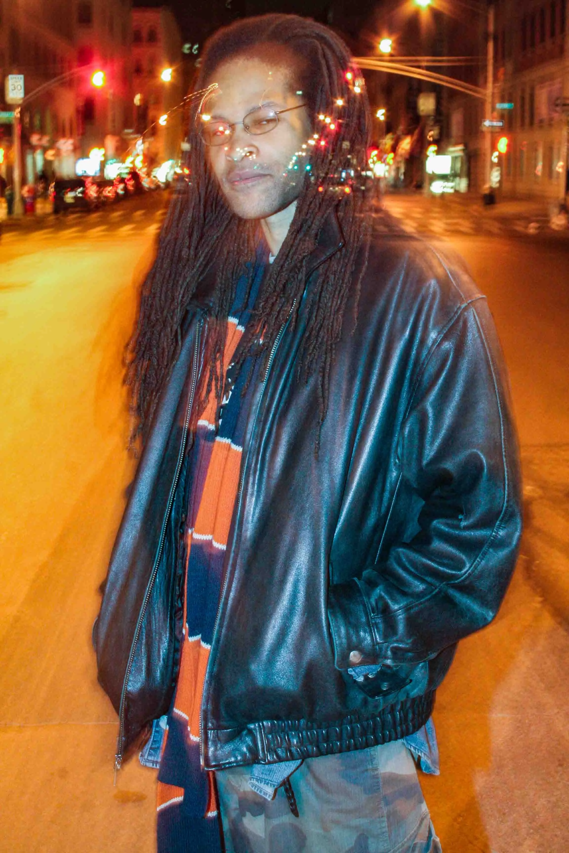A person with dreadlocks and glasses standing on a city street at night. The person is wearing a black leather jacket, a colorful striped scarf, and gray camouflage pants. The street is illuminated by streetlights and vehicle lights, with buildings a