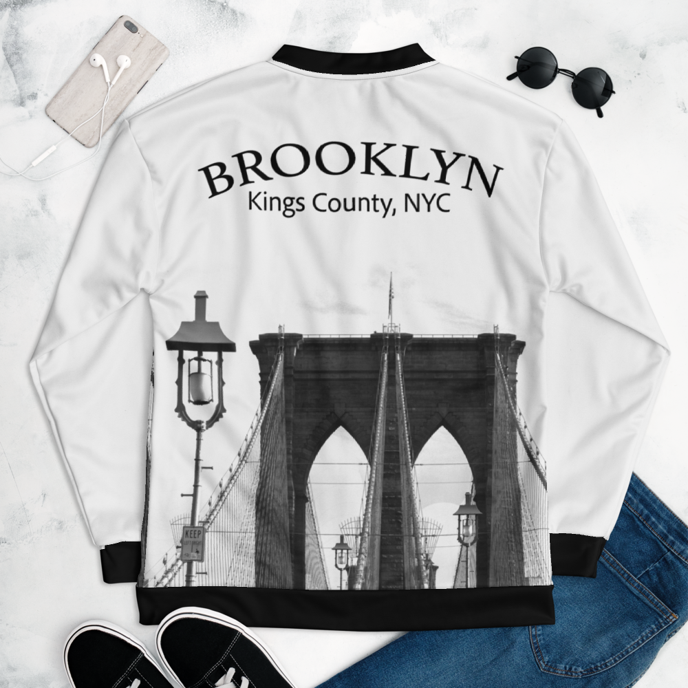 White and black Brooklyn sweatshirt with a black and white image of the Brooklyn Bridge. Also visible are a pair of sunglasses, a pair of black shoes, a pair of blue jeans, a smartphone with earphones, on a white textured background.
