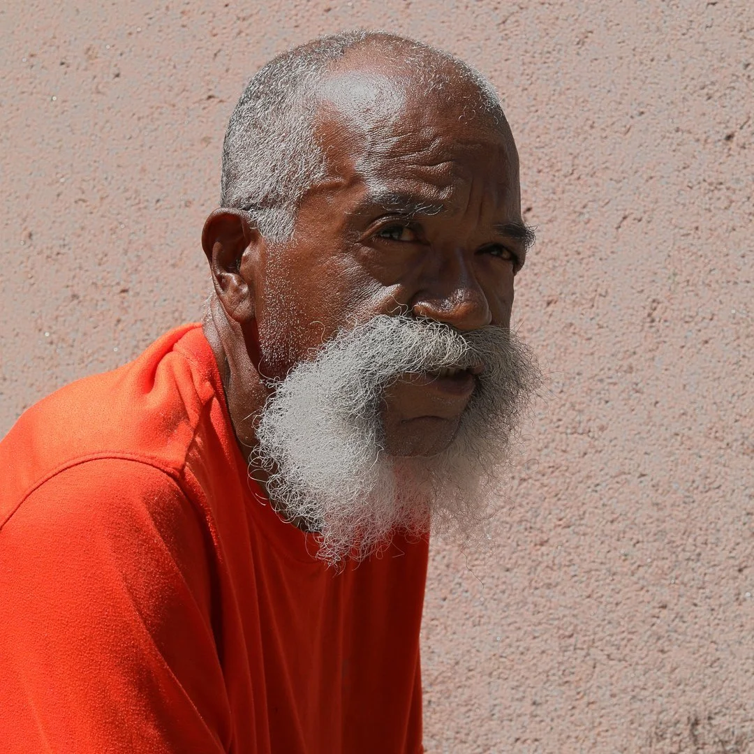 sits outside in downtown, St. Louis, with his white beard and long white mustache, dressed in a bright orange shirt, gazes thoughtfully into the distance. The background is a simple, neutral surface, highlighting the contrast.