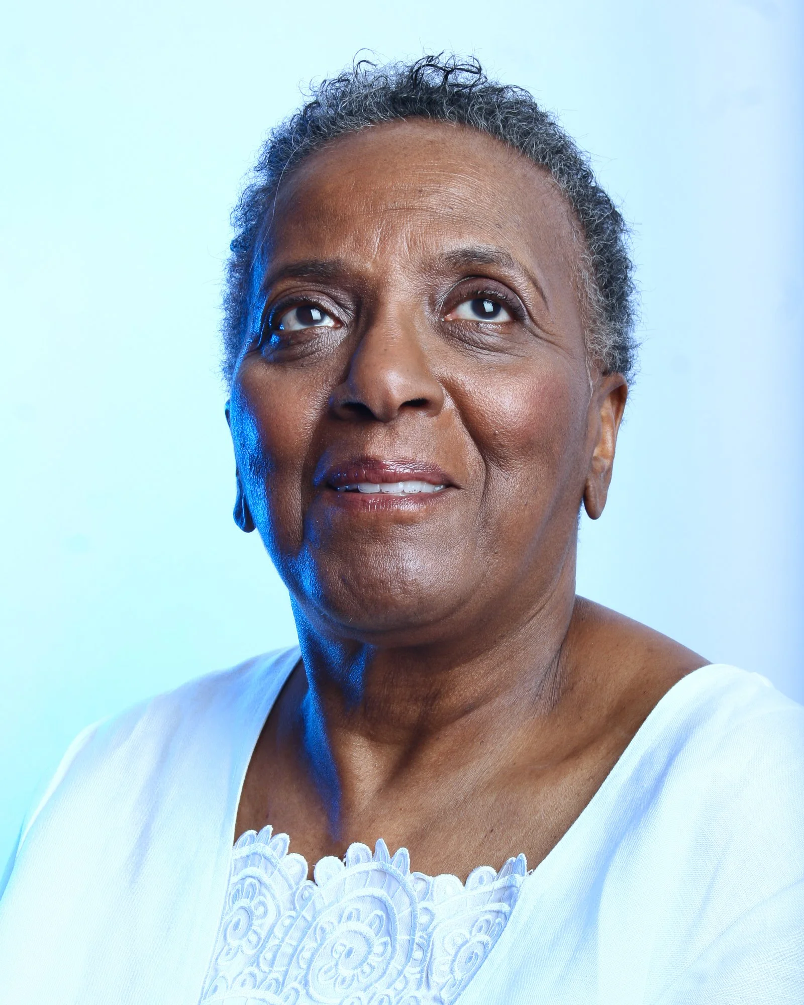 Close-up of an African American woman with short, curly gray hair, wearing a light blue top with white lace detail, against a plain light blue background.