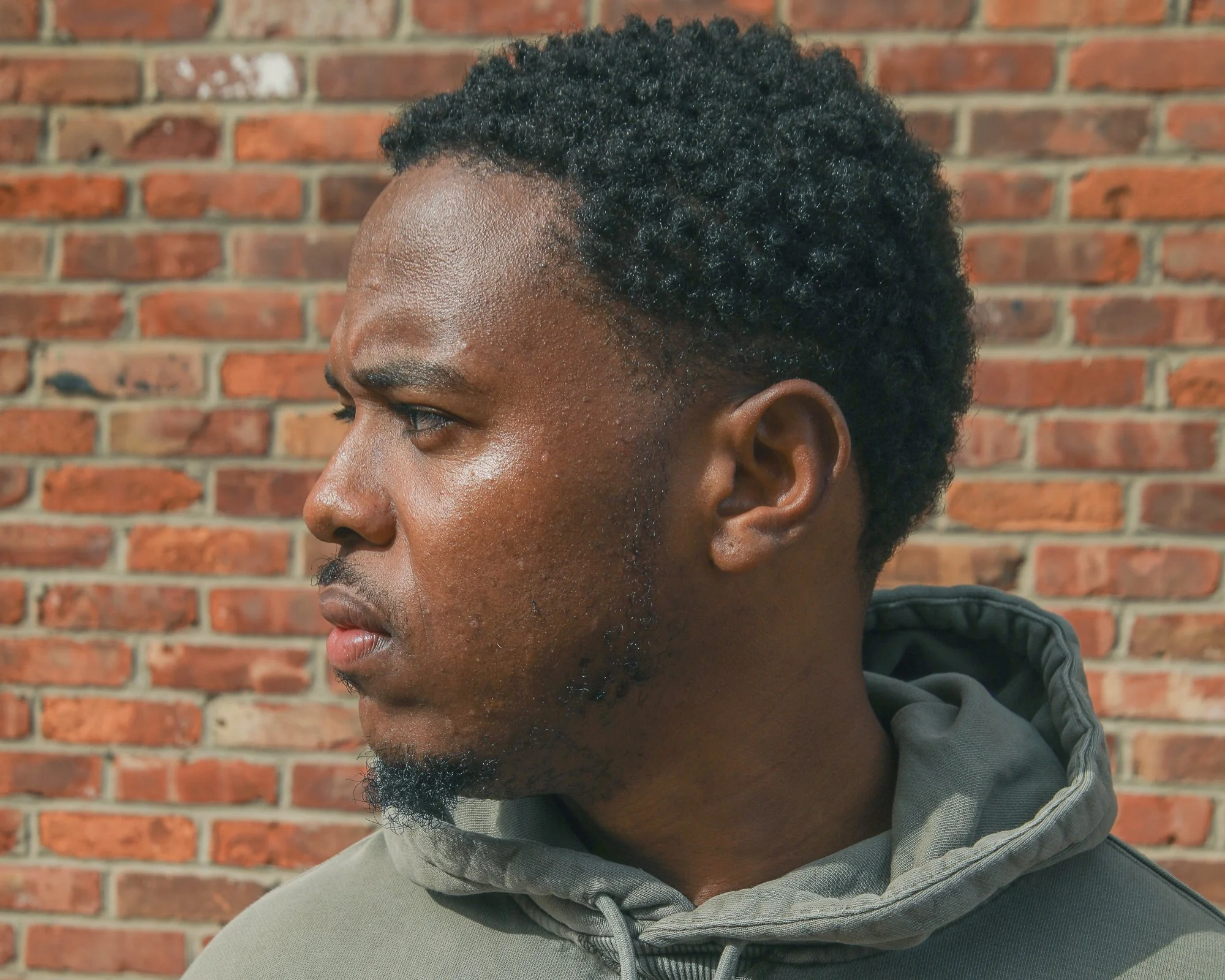 Side profile view of a young Black man with short curly hair, a mustache, and a beard, wearing a gray hoodie, standing in front of a red brick wall.