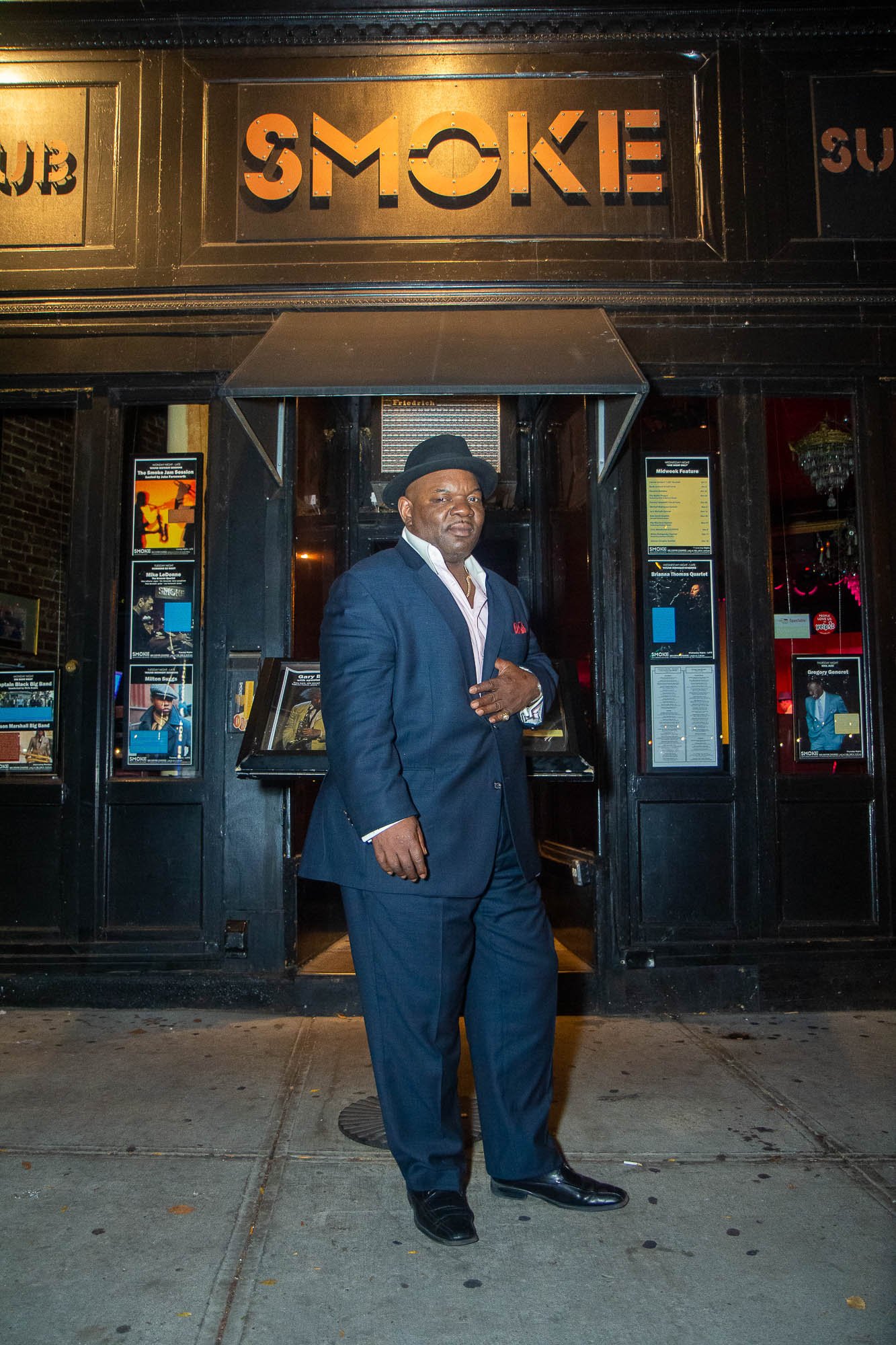 In the captivating image showcasing Stevie Williams's confident demeanor outside the iconic "Smoke Jazz and Supper Club" in Harlem, his poised stance exudes style and sophistication. Adorned in a sleek blue suit, complemented by a fashionable hat and