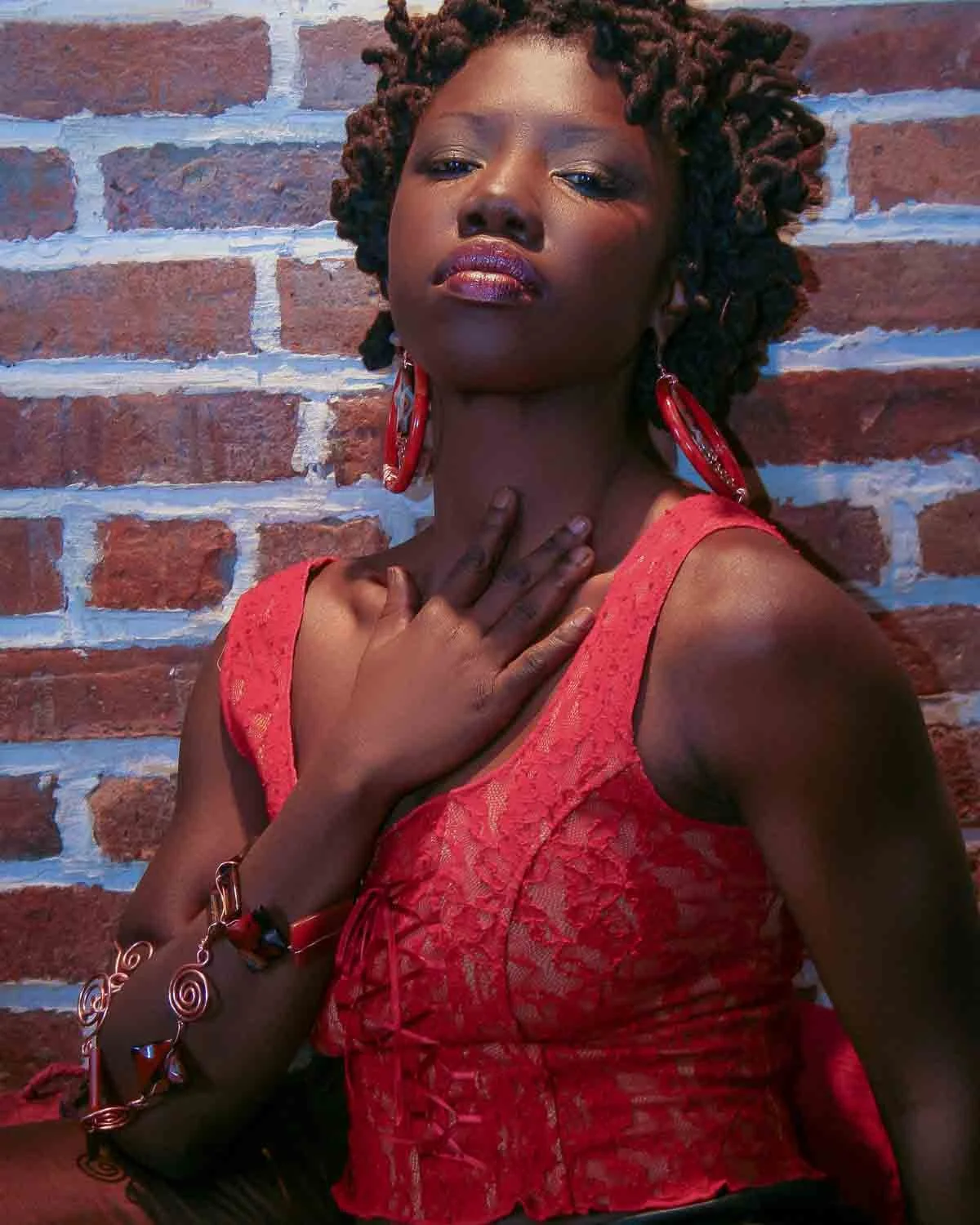 A woman with curly braids, wearing a red lace dress and red earrings, posing against a brick wall with her hand on her neck.
