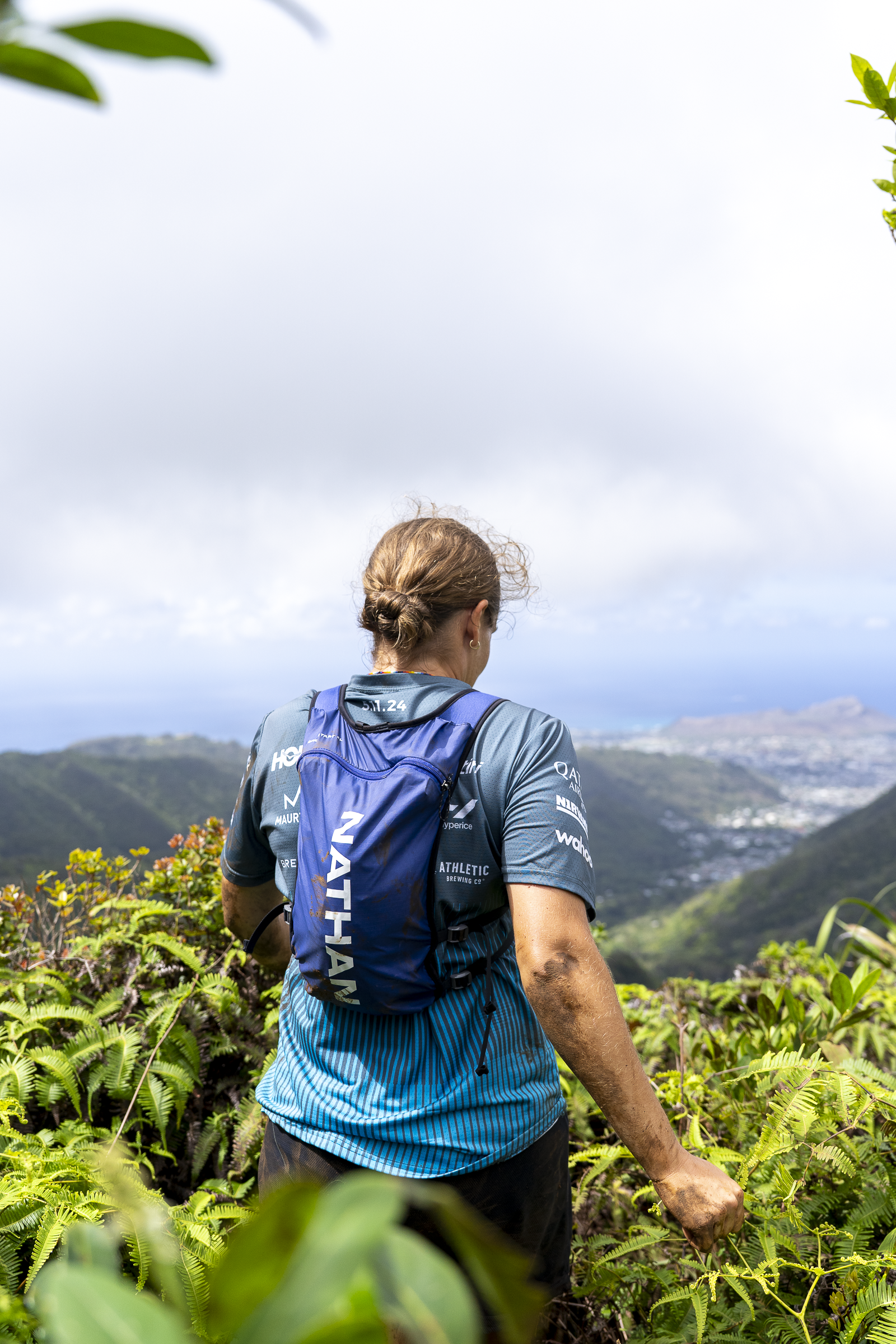 A woman with curly hair tied back, wearing a blue Nike backpack and athletic shirt, hiking through lush green foliage with mountainous landscape and cloudy sky in the background.