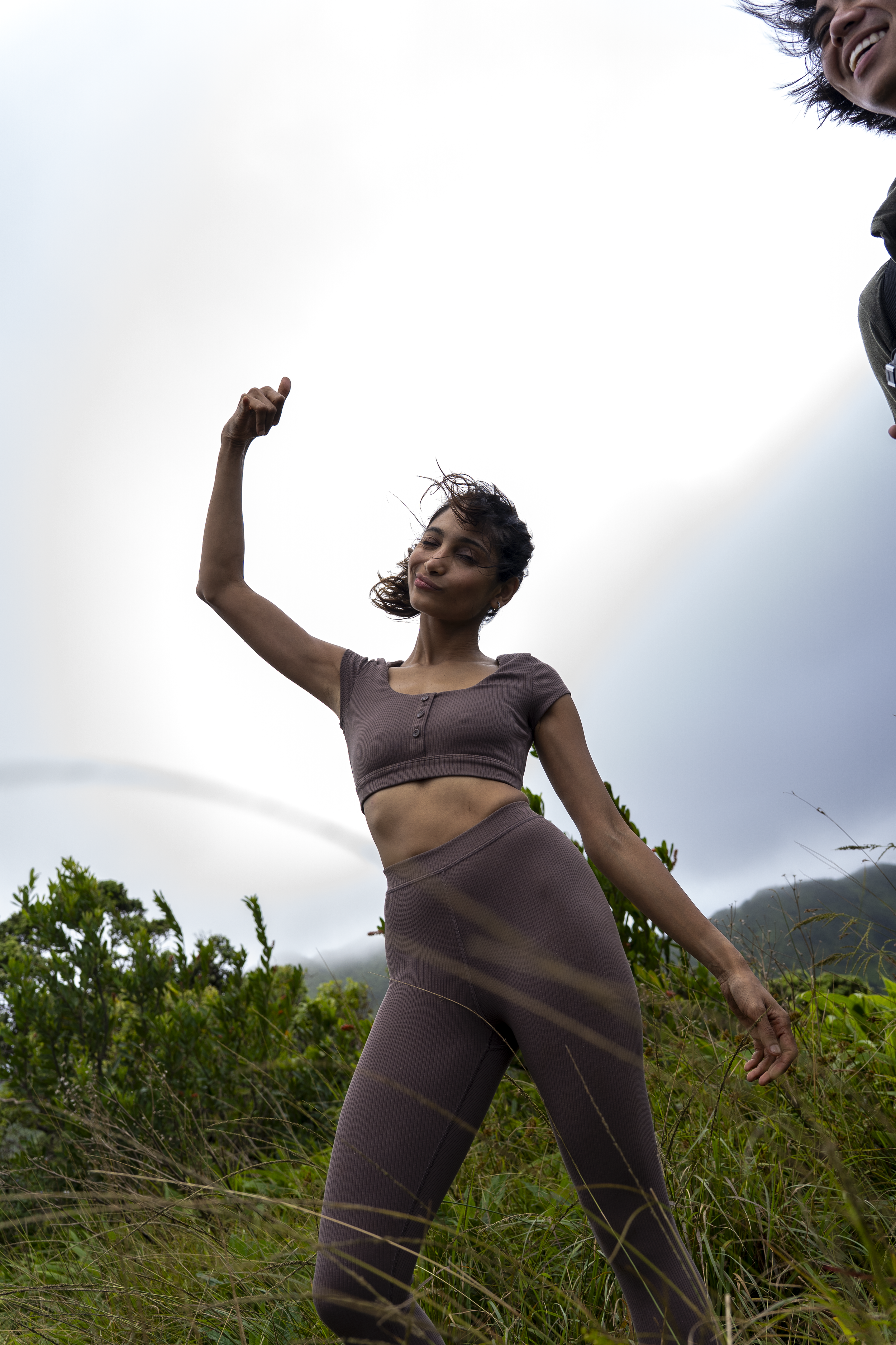 A woman standing in a grassy field with cloudy sky background, flexing her arm and smiling, wearing a matching crop top and leggings in brown.