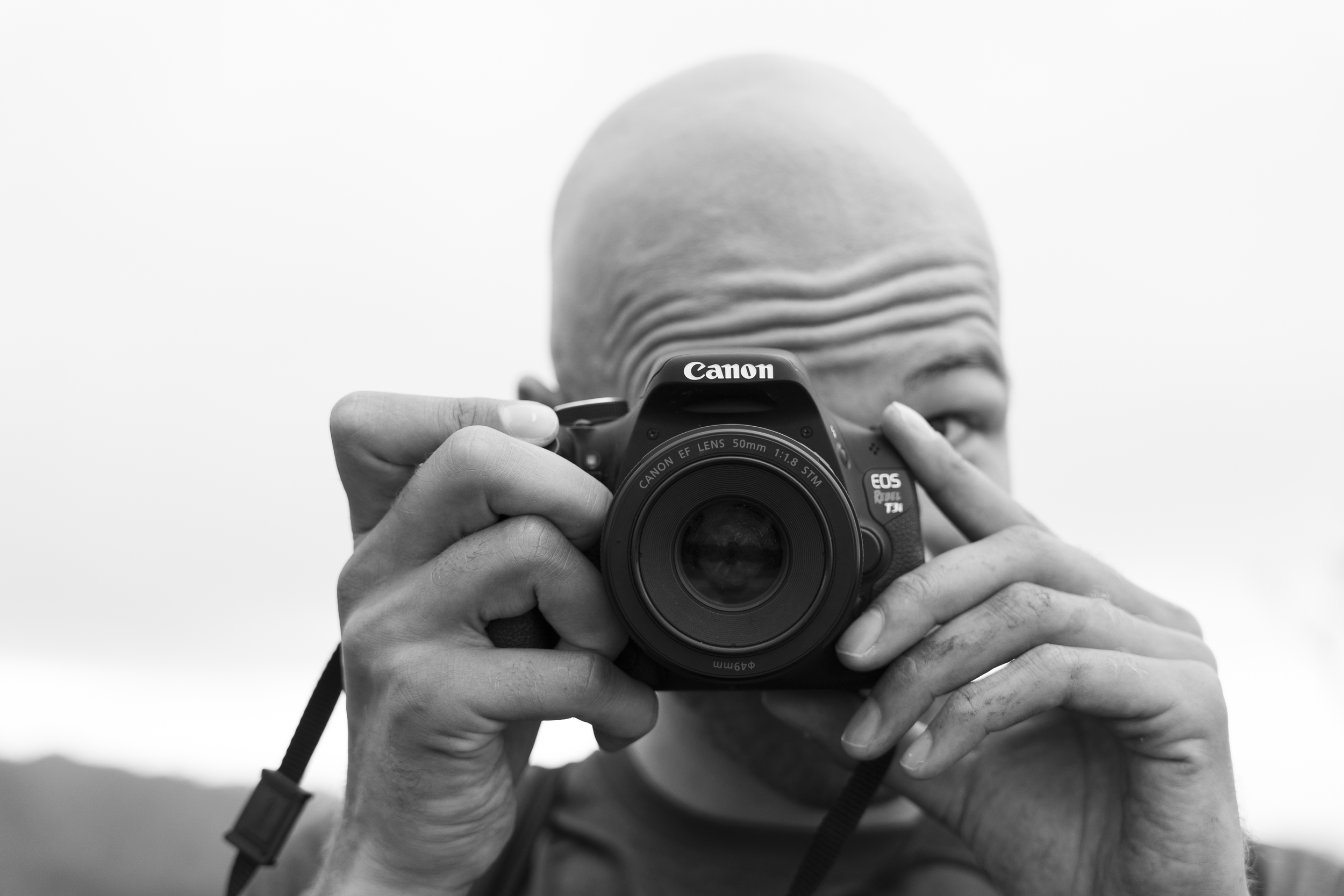 A person with a bald head holding a Canon DSLR camera up to their face, aiming it directly at the viewer.