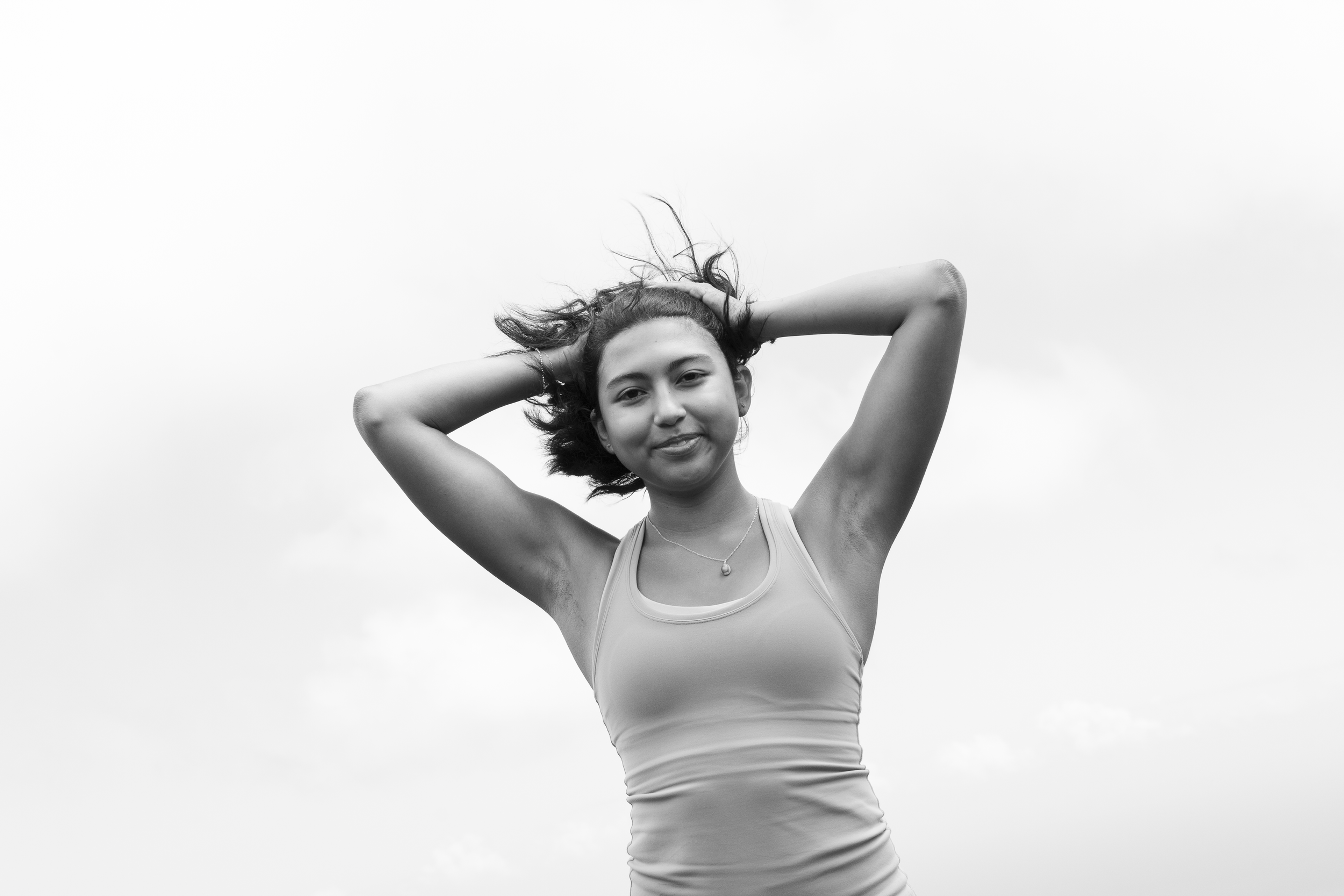 A young woman with short hair smiling, standing outdoors with her hands behind her head, wearing a tank top and a necklace, against a cloudy sky