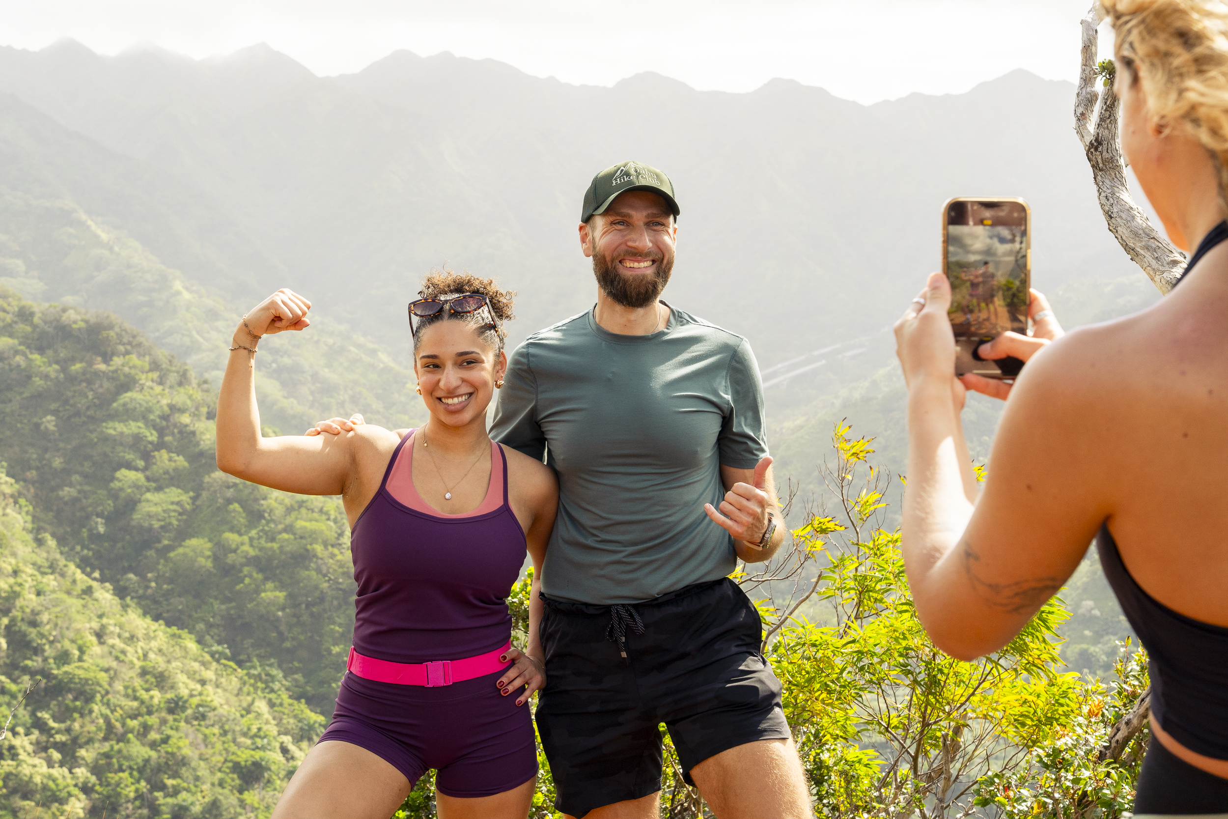 Hikers enjoying a hike in a mountainous area. They are taking a selfie, with one woman flexing her arm and smiling at the camera, and a man smiling proudly. The background features lush green hills and a misty mountain range.