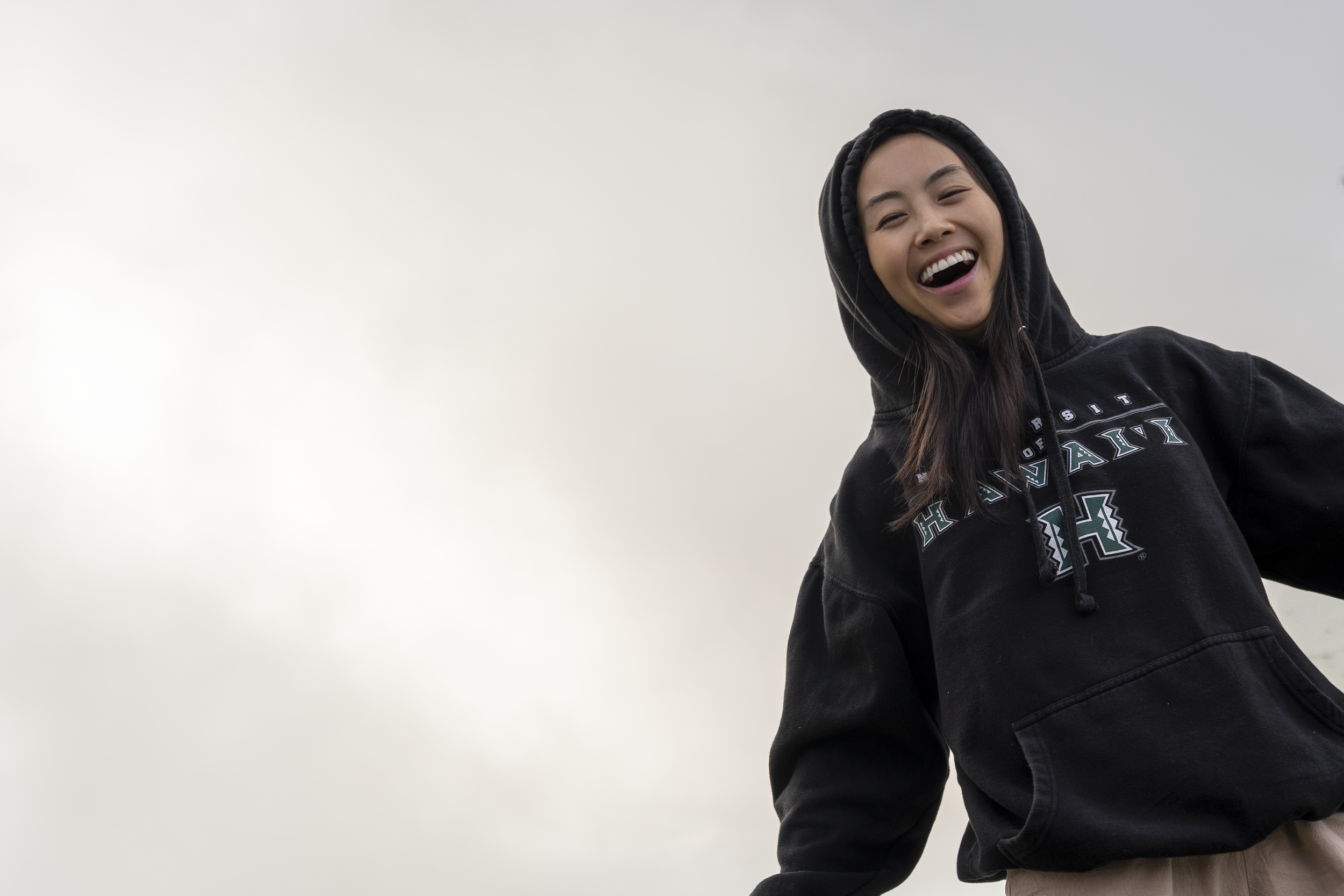 A young woman wearing a black hoodie with the words Hawaii and an H logo, smiling and laughing against a cloudy sky.