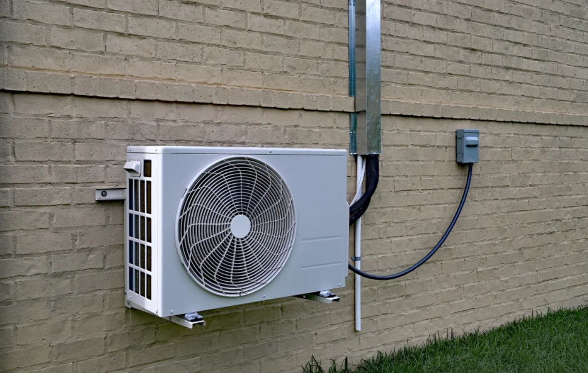 Outdoor unit of a heat pump mounted on a textured beige brick wall, with a visible fan and connected piping.