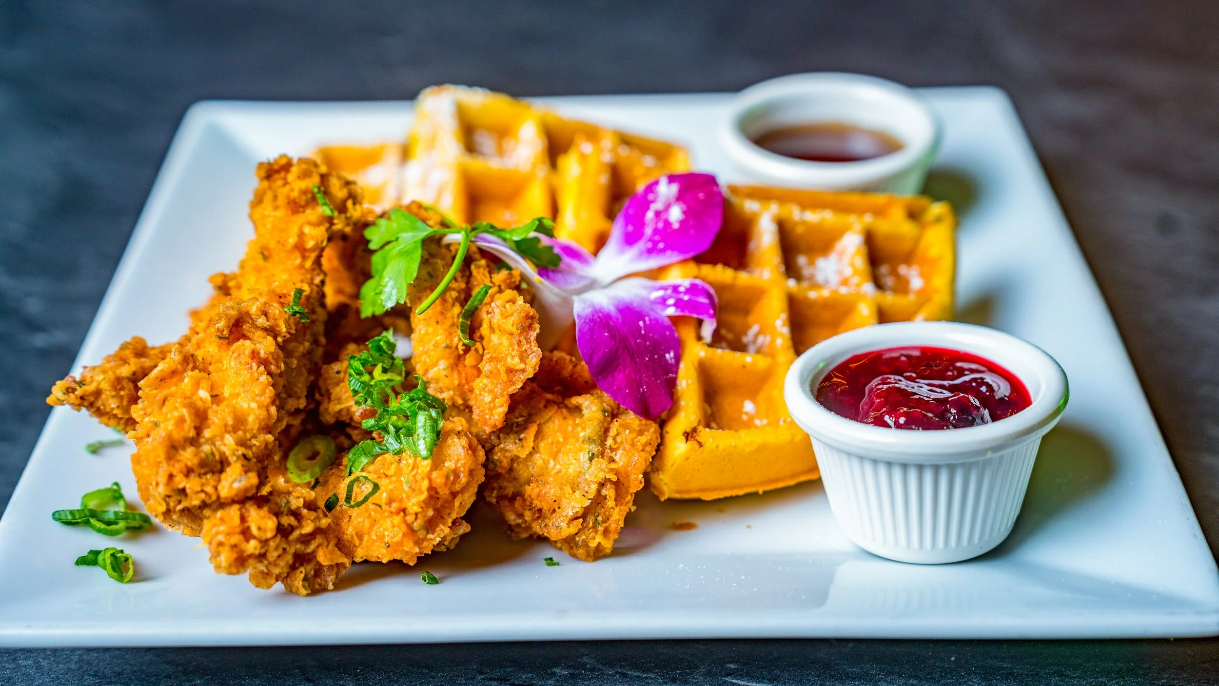 Best brand food photography waffles with powdered sugar topped with fried chicken tenders orchid flower and herbs served with syrup and berry jam on dark surface in New York City and shot by Lumumba David from David Media Group