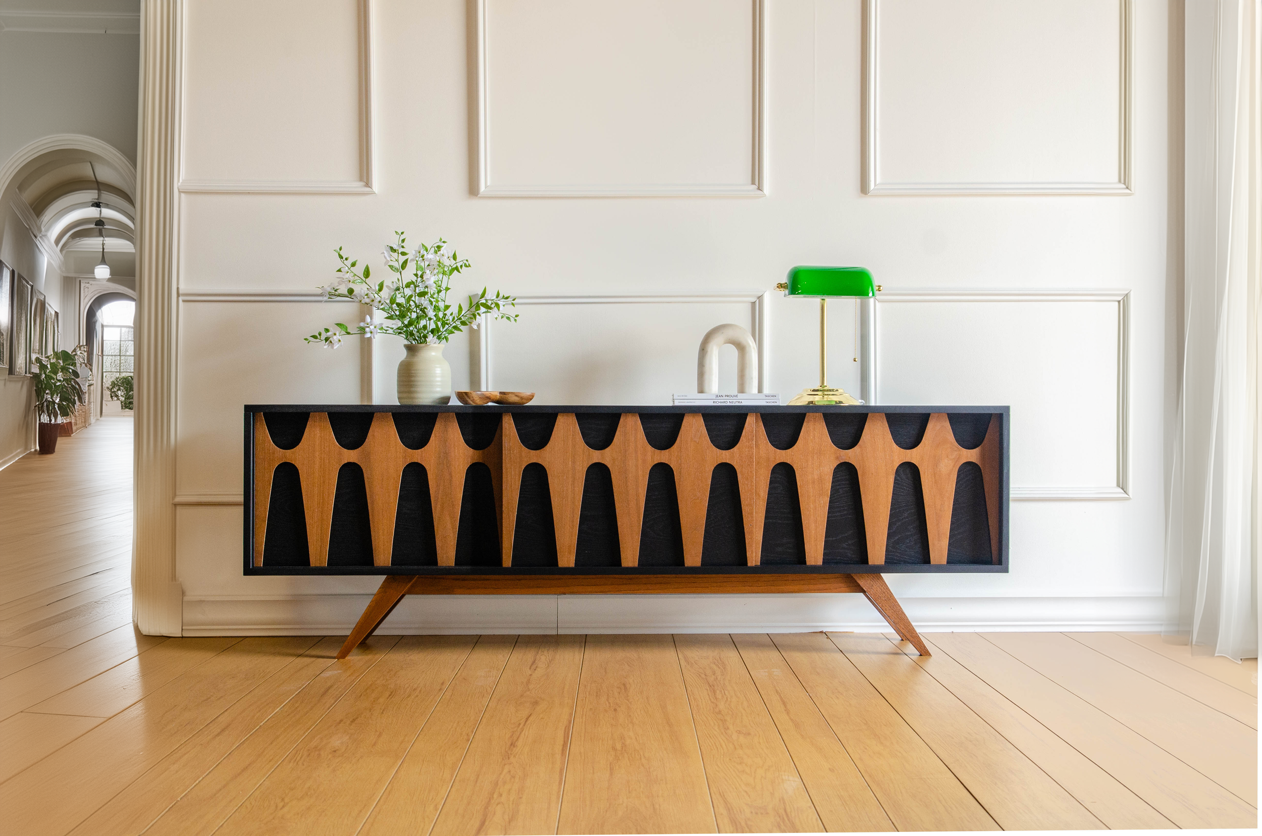 A modern wooden sideboard with black patterns on the front, positioned against a white paneled wall in a bright hallway with wooden flooring. On top of the sideboard are decorative items including a green desk lamp, a white sculpture, a vase with green leaves, and books.