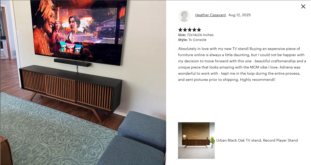 A modern living room with a large flat-screen TV mounted on the wall above a black and wood TV stand. The TV is displaying a scene with several people, including a woman with blonde hair and a man in a striped shirt. The room has wood flooring, a patterned rug, and a blue upholstered chair in the foreground.