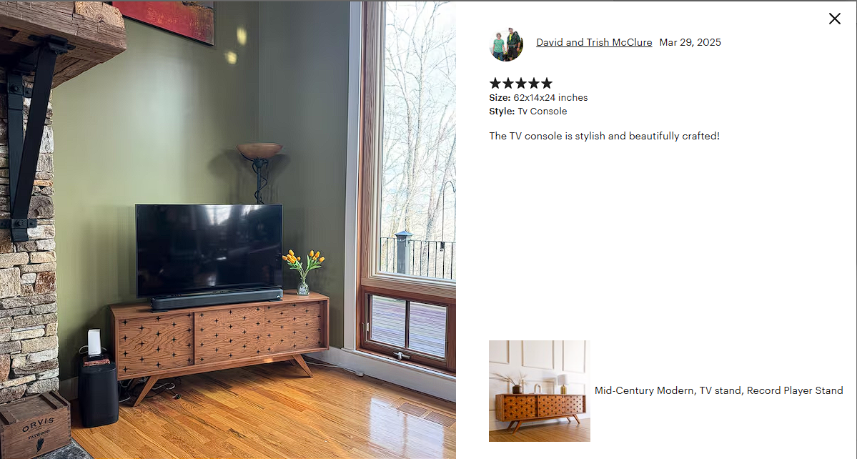 Mid-century modern wooden TV stand with a flat-screen TV, a small white speaker, a vase with yellow flowers, and a floor lamp in a room with hardwood floors and large window showing trees outside.