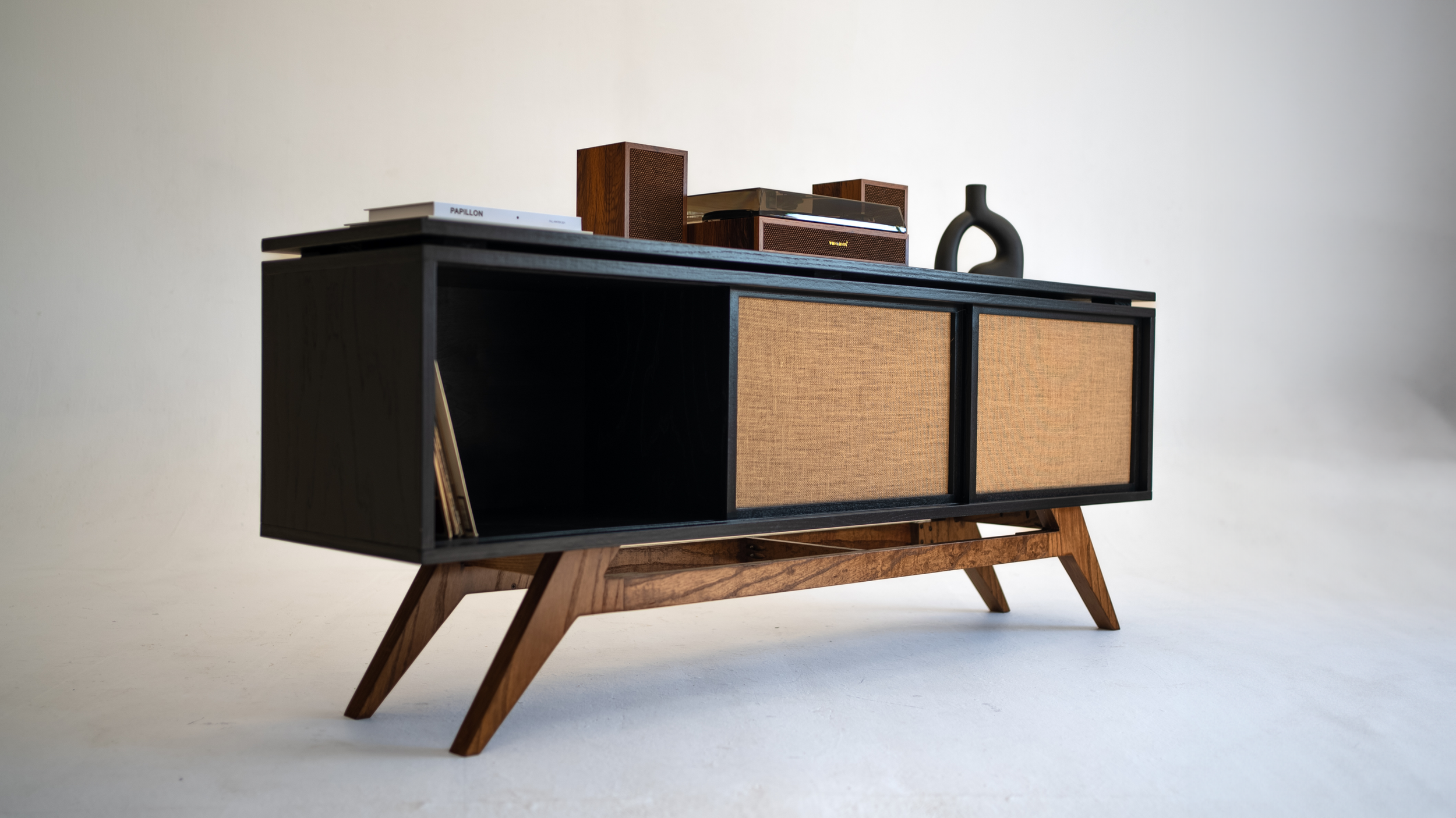 Mid-century modern black and wooden sideboard with two sliding doors with woven rattan panels, decorated with vintage stereo speakers, a black sculptural vase, books, and a record player on top.